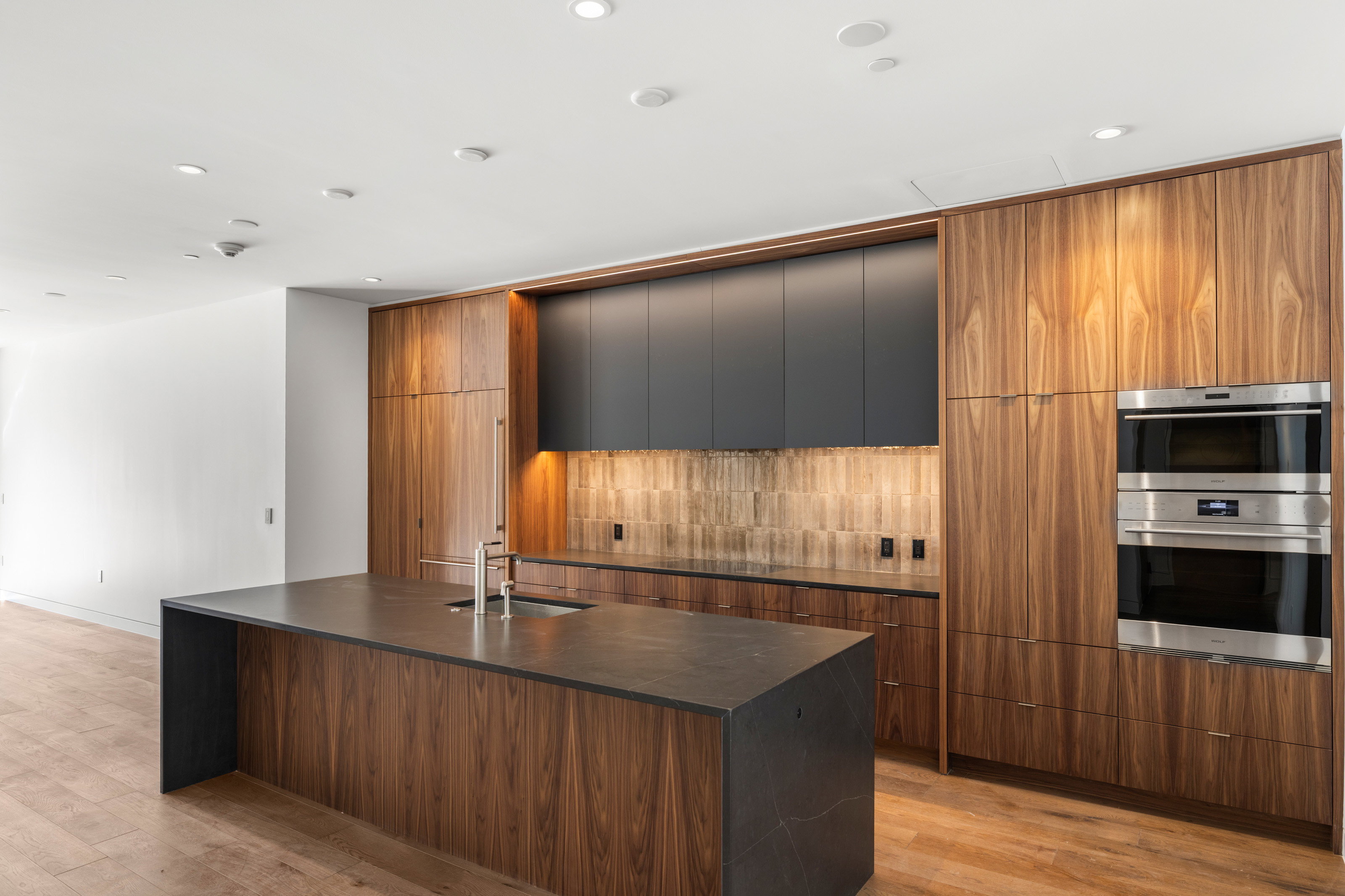 Gourmet kitchen featuring a large central island with an undermount sink, integrated stainless steel appliances including double ovens, full-height wood-finish cabinetry, and contemporary dark matte upper cabinets
