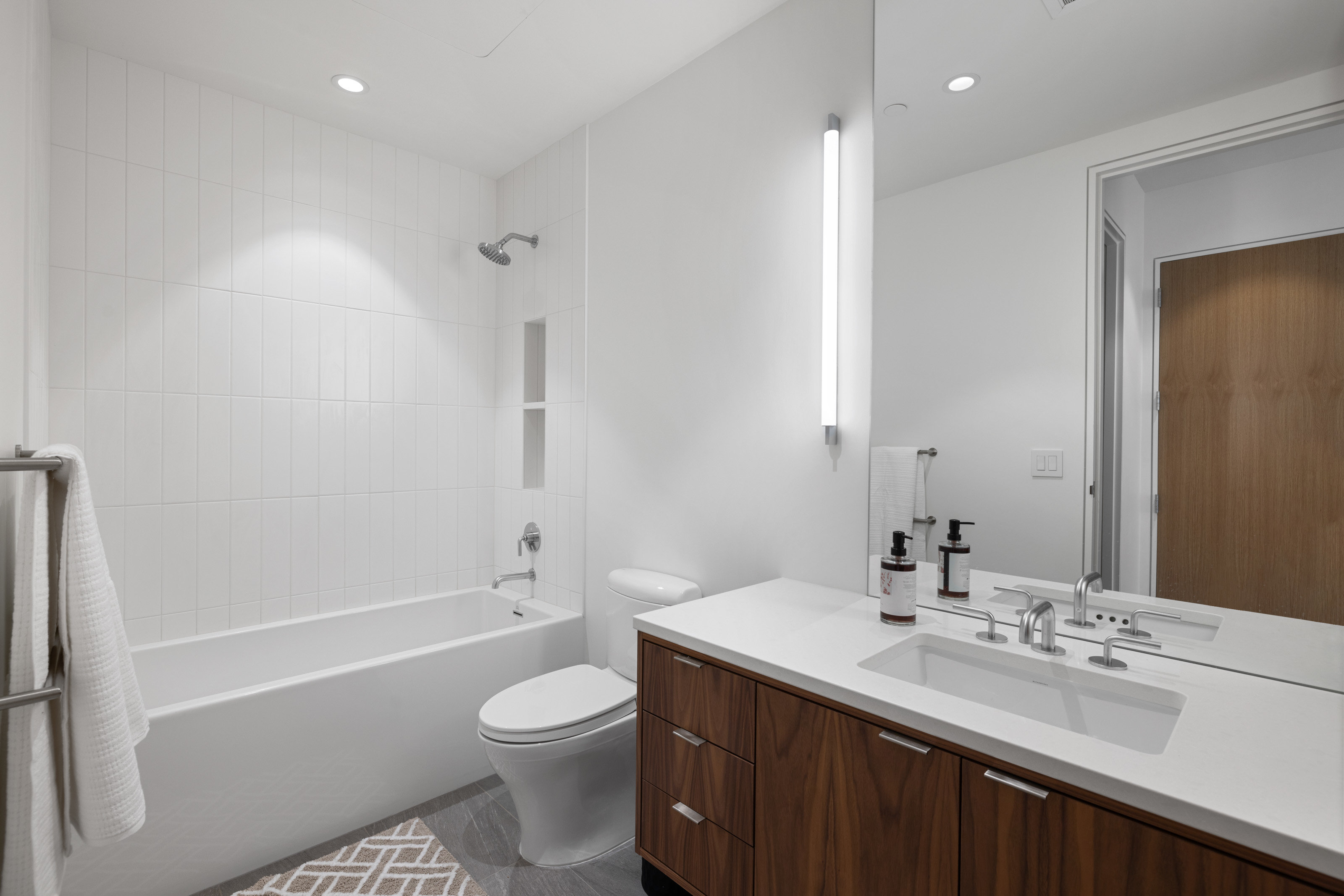 Contemporary bathroom featuring a white subway tile tub surround, built-in shower niche, wood-finish vanity with an integrated sink, and an illuminated vertical mirror