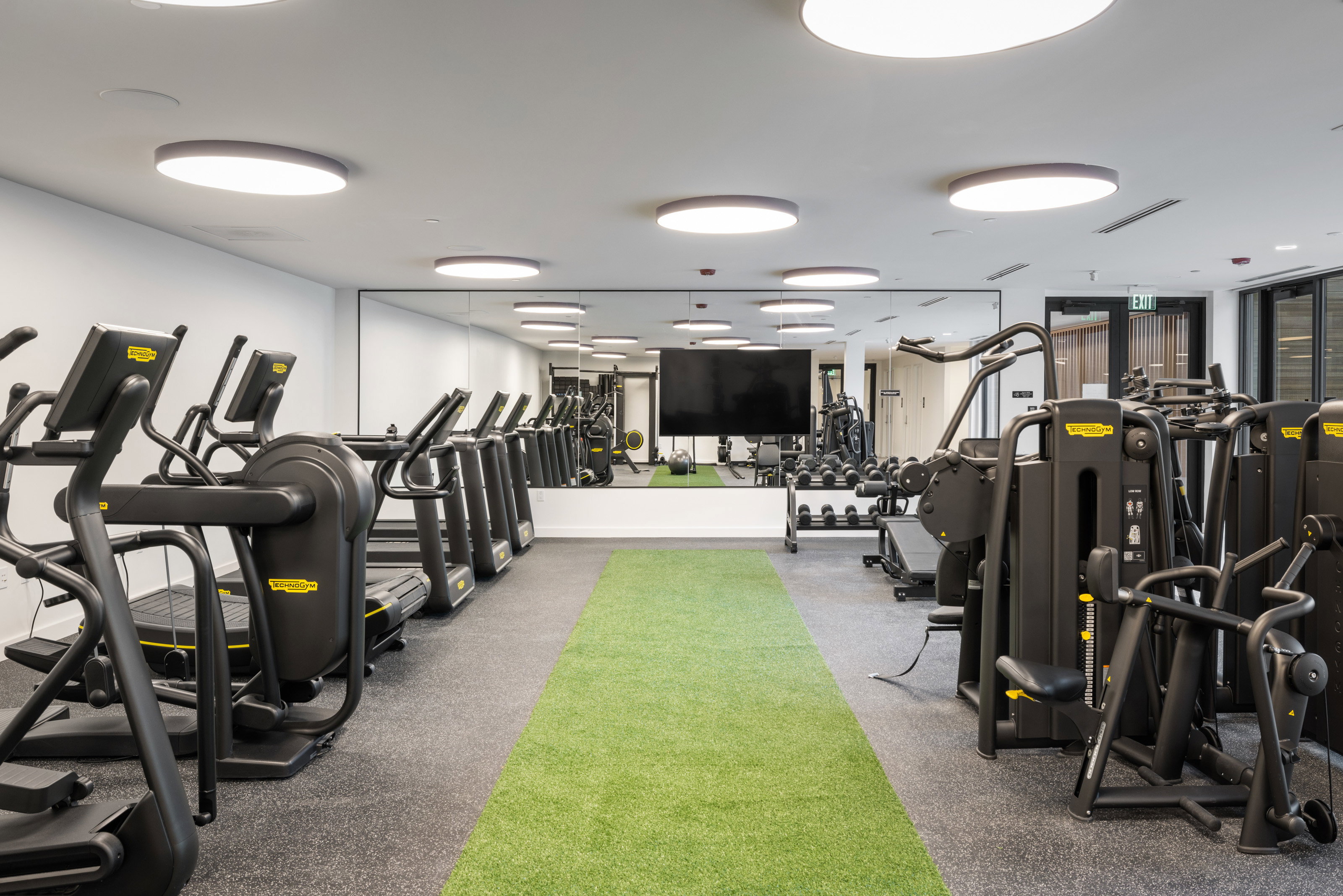 Fitness center featuring a row of cardio equipment, strength training machines, extensive mirrored wall, recessed circular lighting, and a green turf track