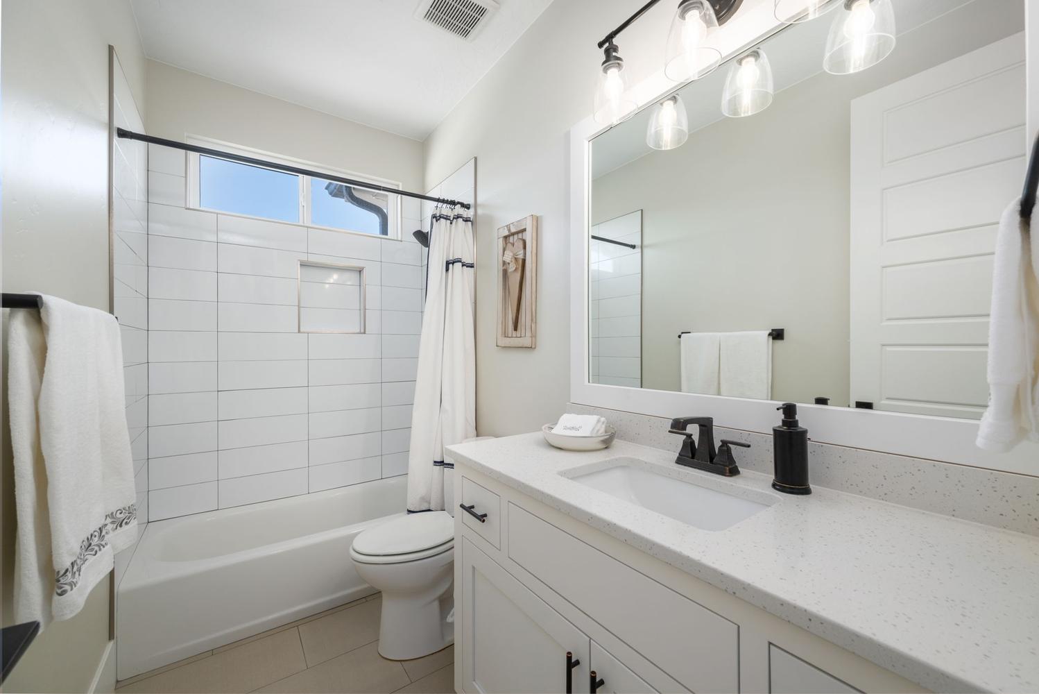 Bathroom featuring a white vanity with an integrated sink and light-colored countertop, matte black fixtures, a framed mirror with overhead lighting, a white subway-tiled tub/shower combination with a built-in niche, and neutral-toned floor tiling