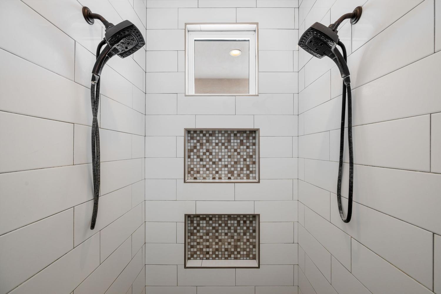Shower with white subway tile, dual dark bronze showerheads, two recessed mosaic tile niches, and an upper transom window