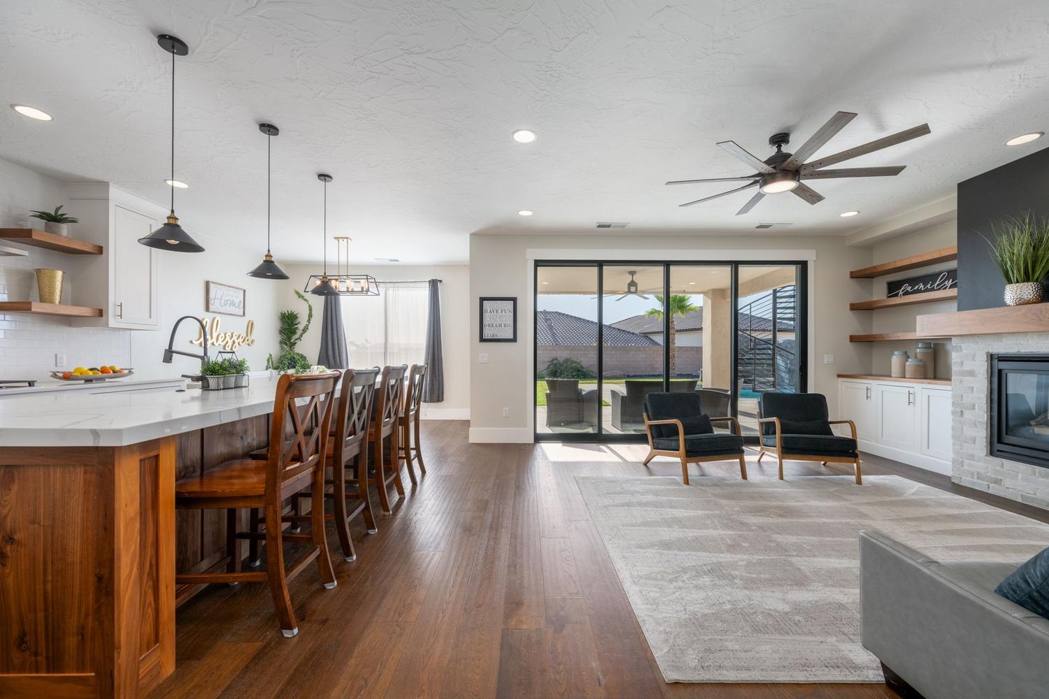 Open-concept living area featuring wood-finish flooring, a kitchen island with seating, a fireplace with a stone surround and wood mantel, built-in shelving, and sliding glass doors to the exterior