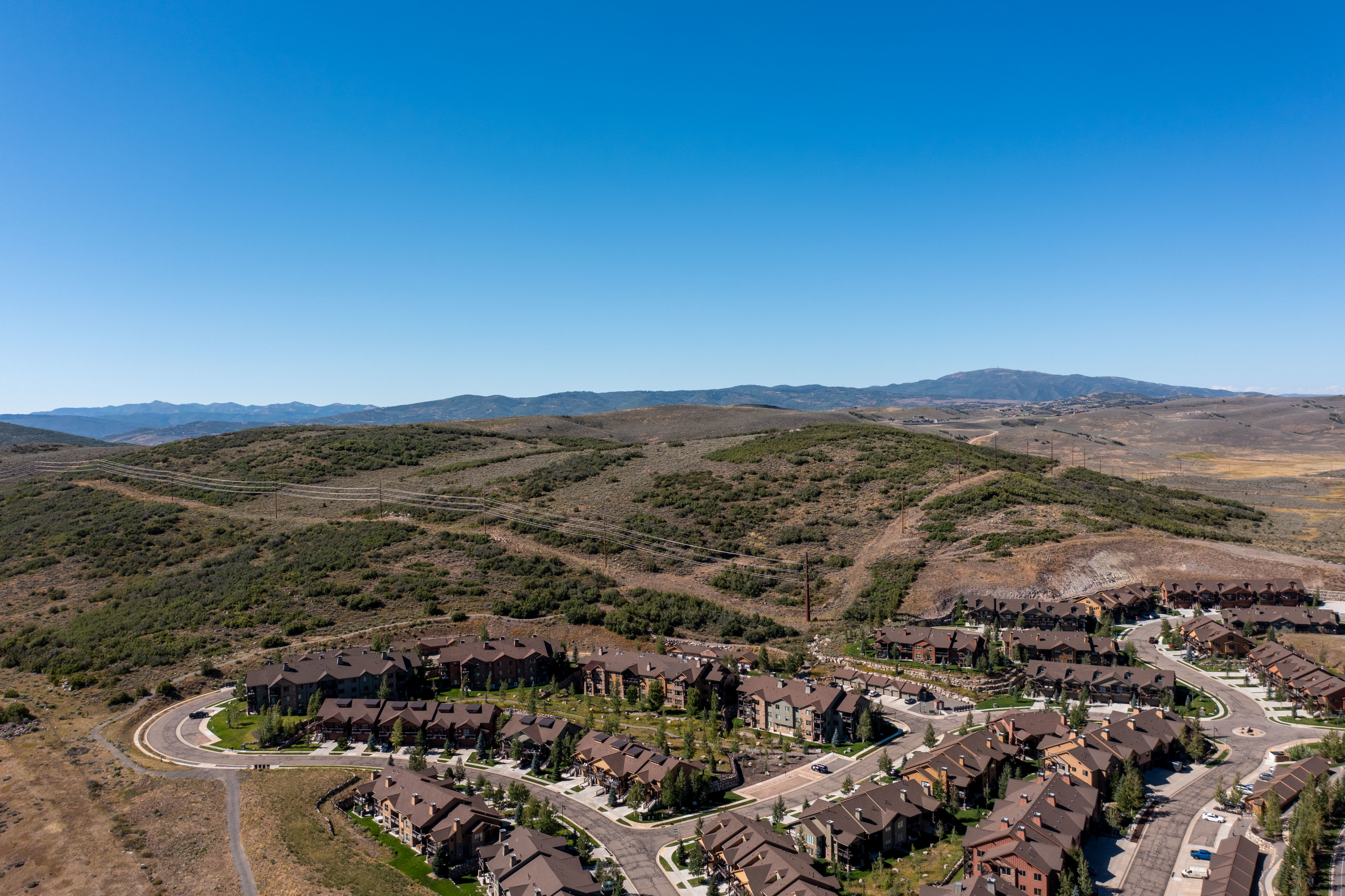 Aerial panorama showcasing a community of properties with dark roofs and varied exterior finishes, surrounded by rolling hills and distant mountains under a clear sky