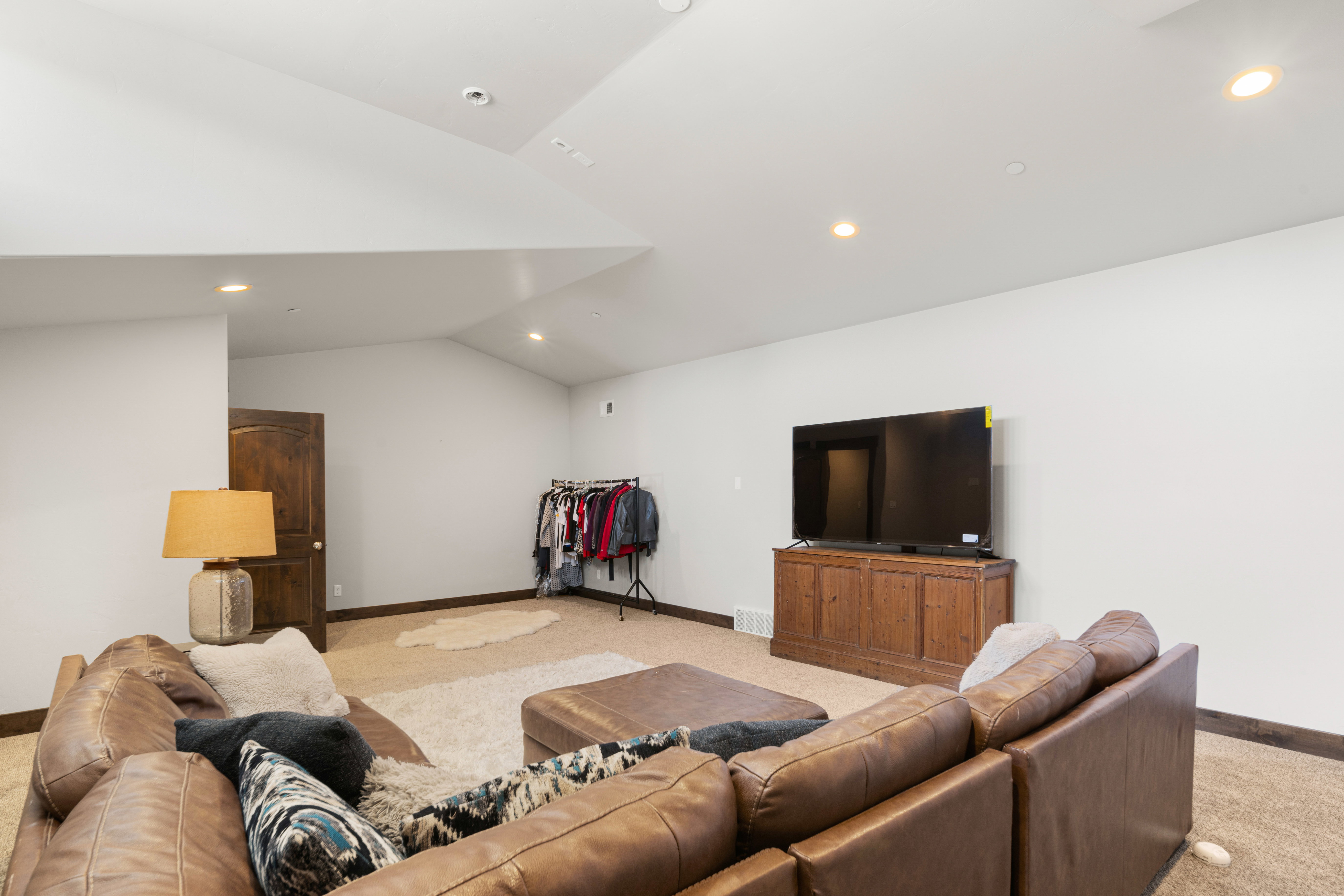 Spacious room featuring vaulted ceilings, recessed lighting, wood-finish baseboards, a dark wood-paneled door, and neutral-toned carpeting