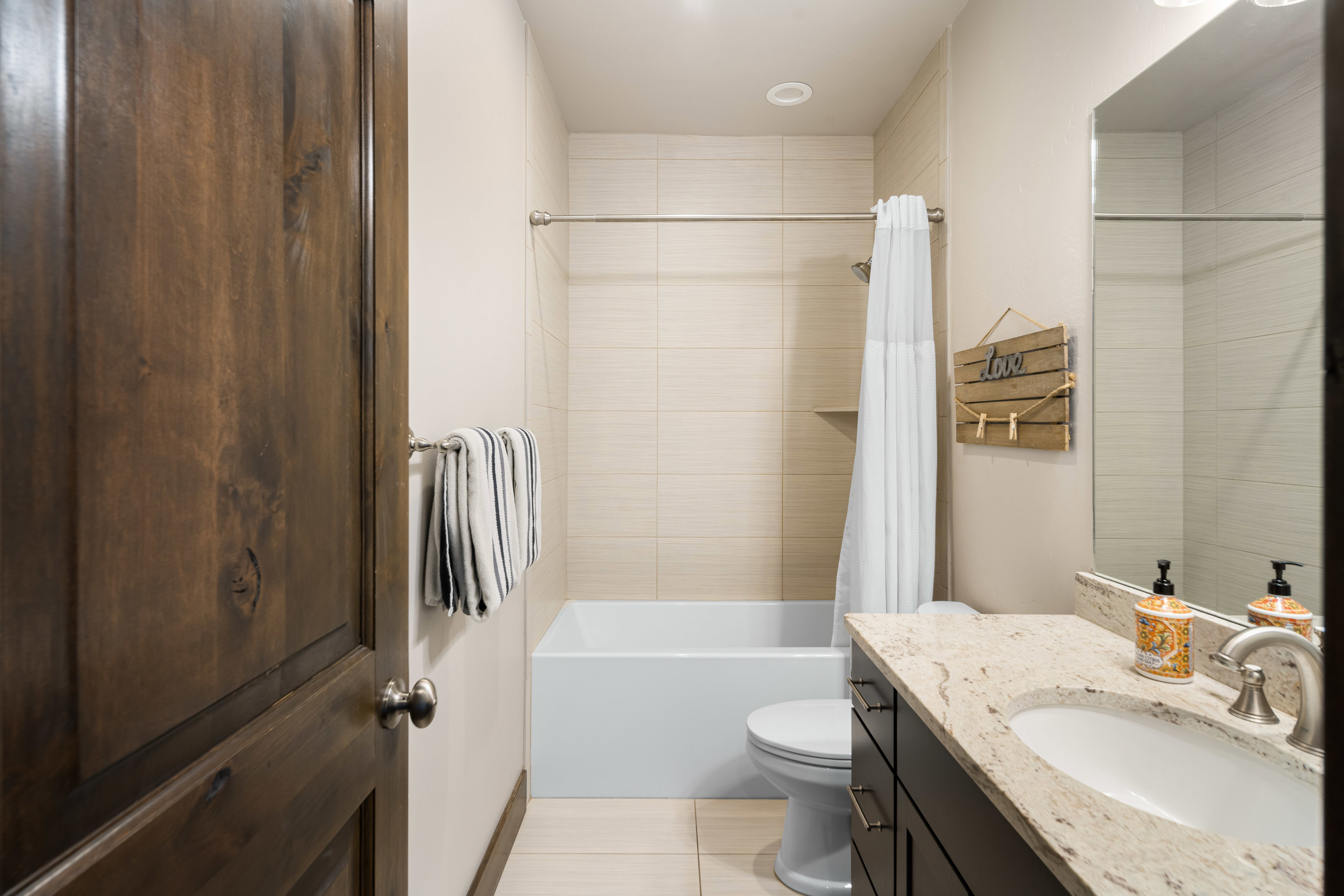Bathroom featuring a white tub with light-toned tiled surround, a dark wood-finish vanity with a light granite countertop, and tile flooring