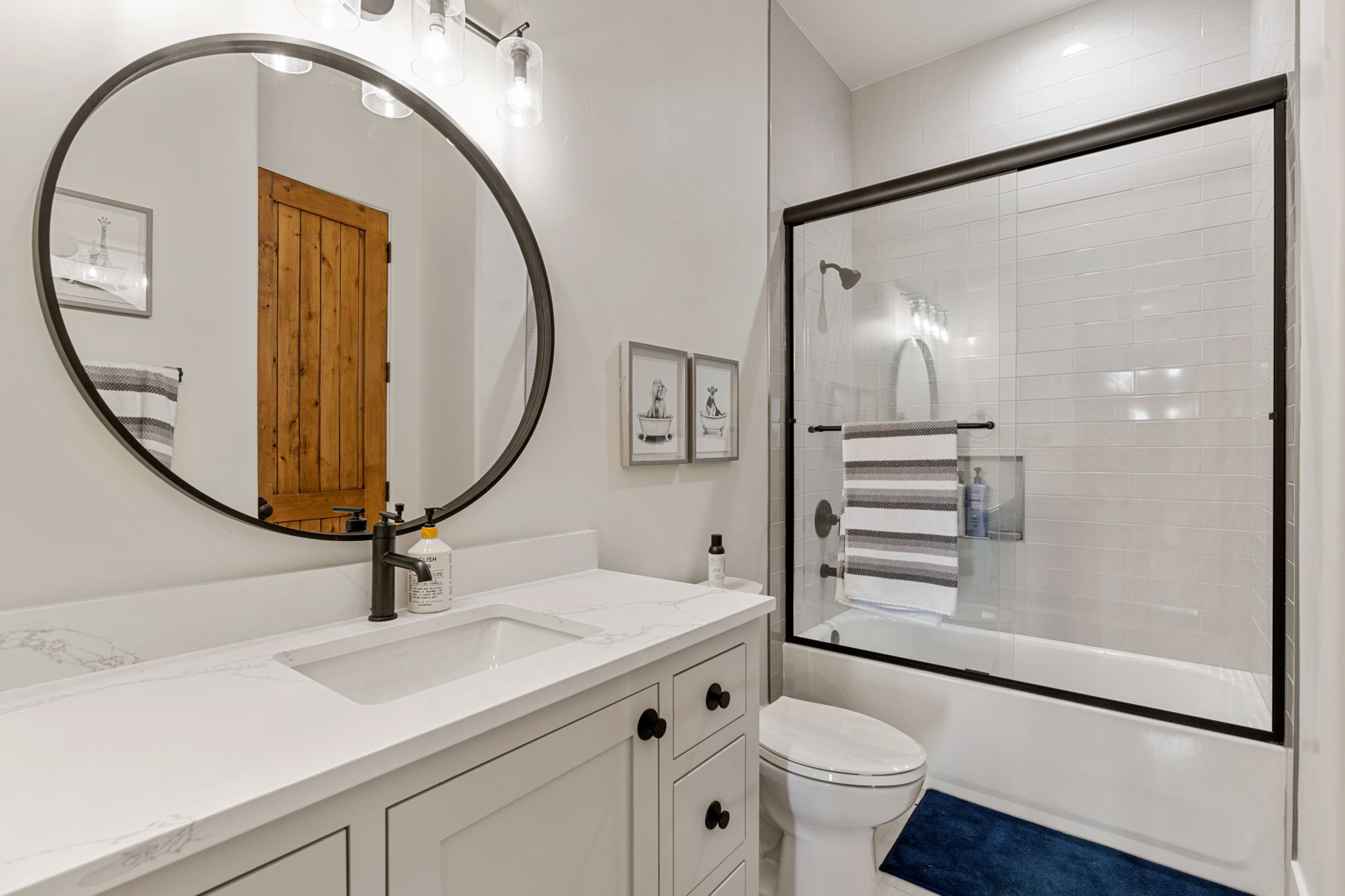 Bathroom featuring a white vanity with an integrated sink, black hardware, a round black-framed mirror, and a tub with a sliding glass shower door