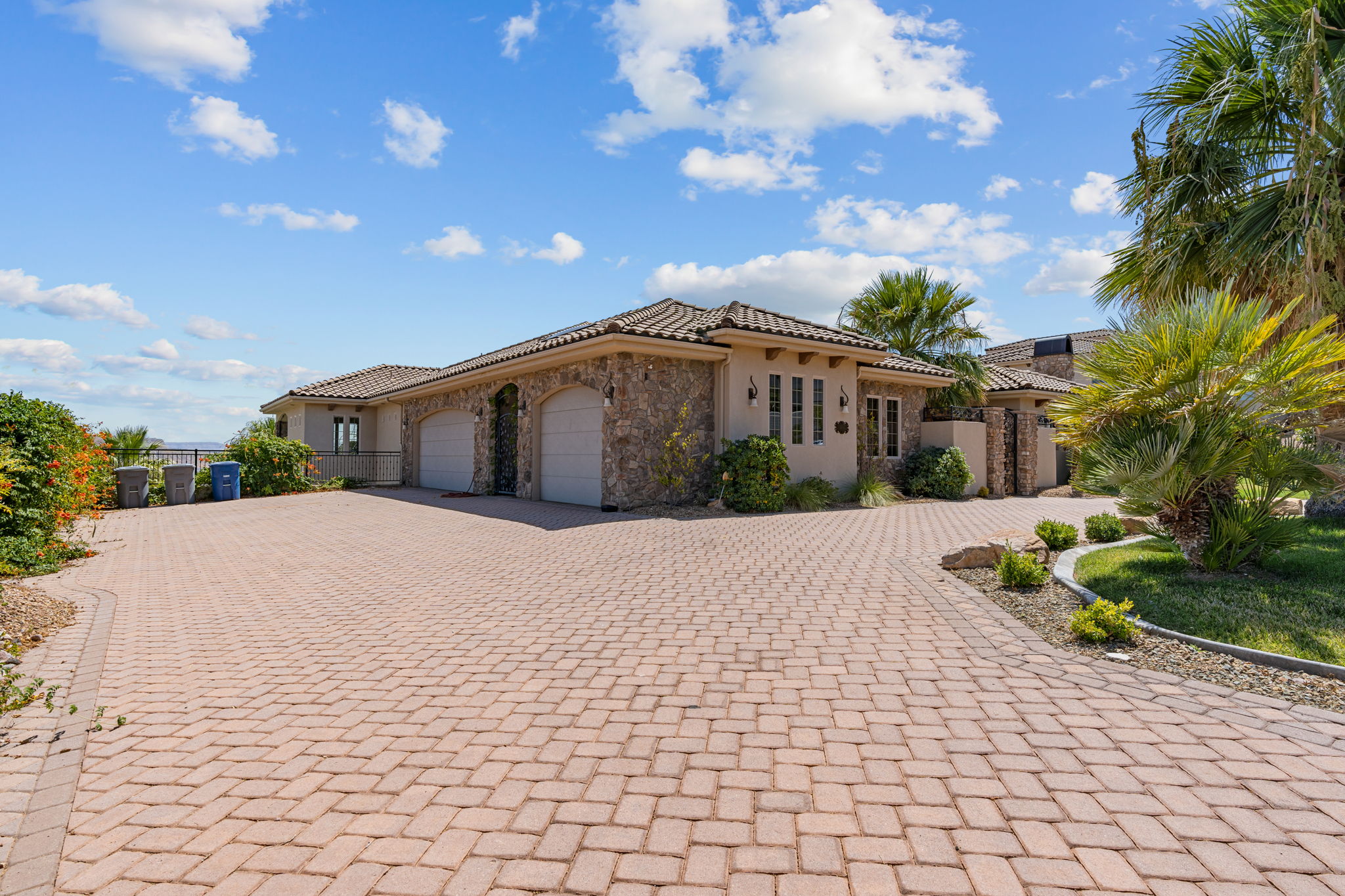 Mediterranean-style architecture with a stone facade, a multi-car garage, a paver driveway, mature palm trees, and lush landscaping