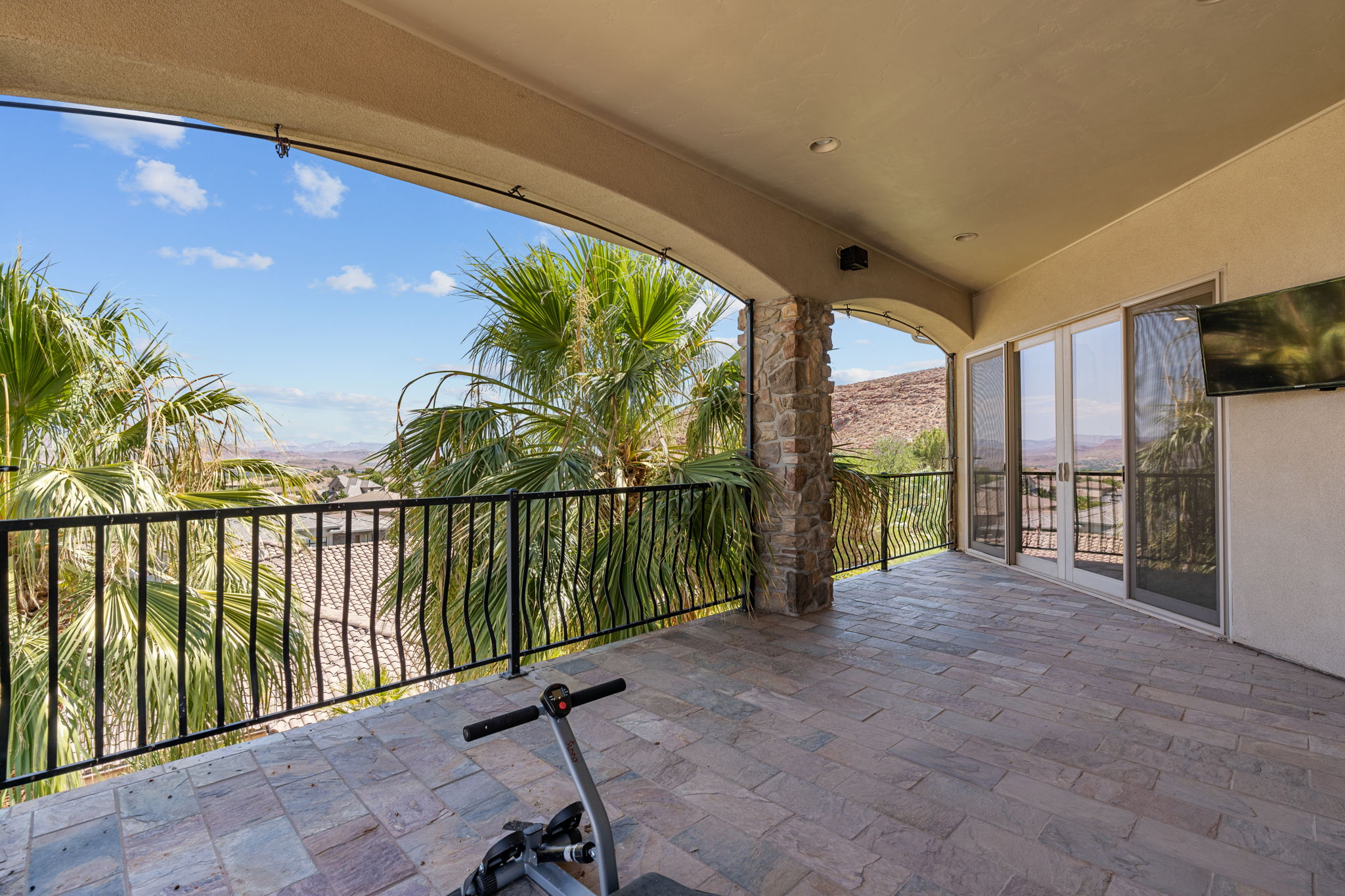 Spacious covered patio featuring stone-look tile flooring, black wrought-iron railing, a stone-clad column, and double glass doors