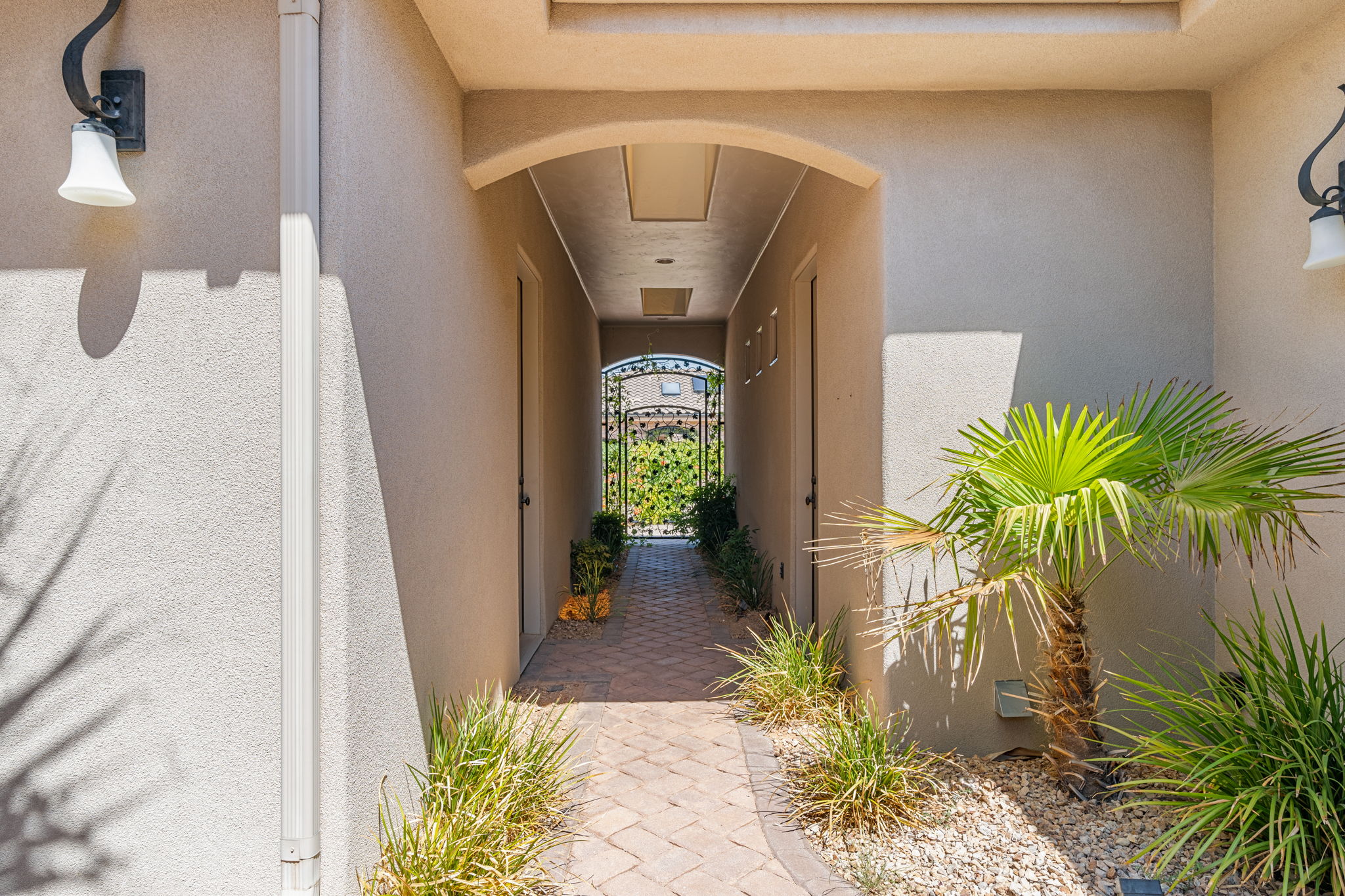 Archway entry corridor featuring stucco exterior, paver walkway, mature landscaping, and an ornate wrought iron gate