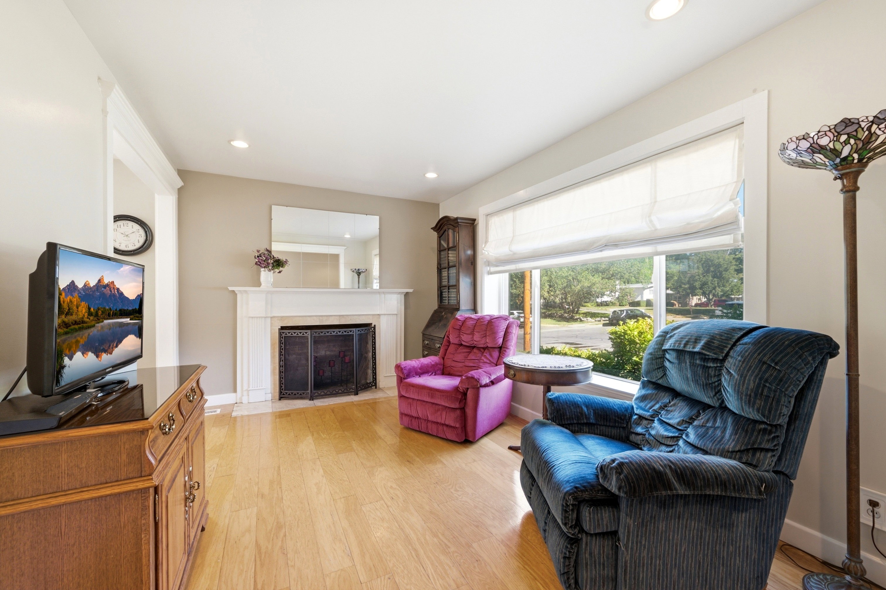 Living area featuring light wood-finish flooring, a fireplace with a white mantel and hearth, and a large window with privacy blinds