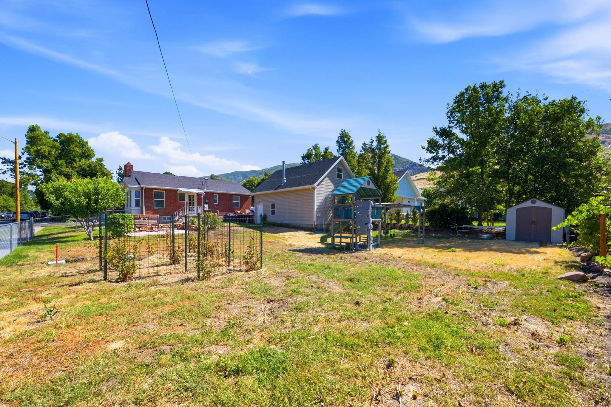 Expansive backyard featuring a red brick dwelling with a patio, a detached beige accessory structure, a fenced garden area, and a metal storage shed