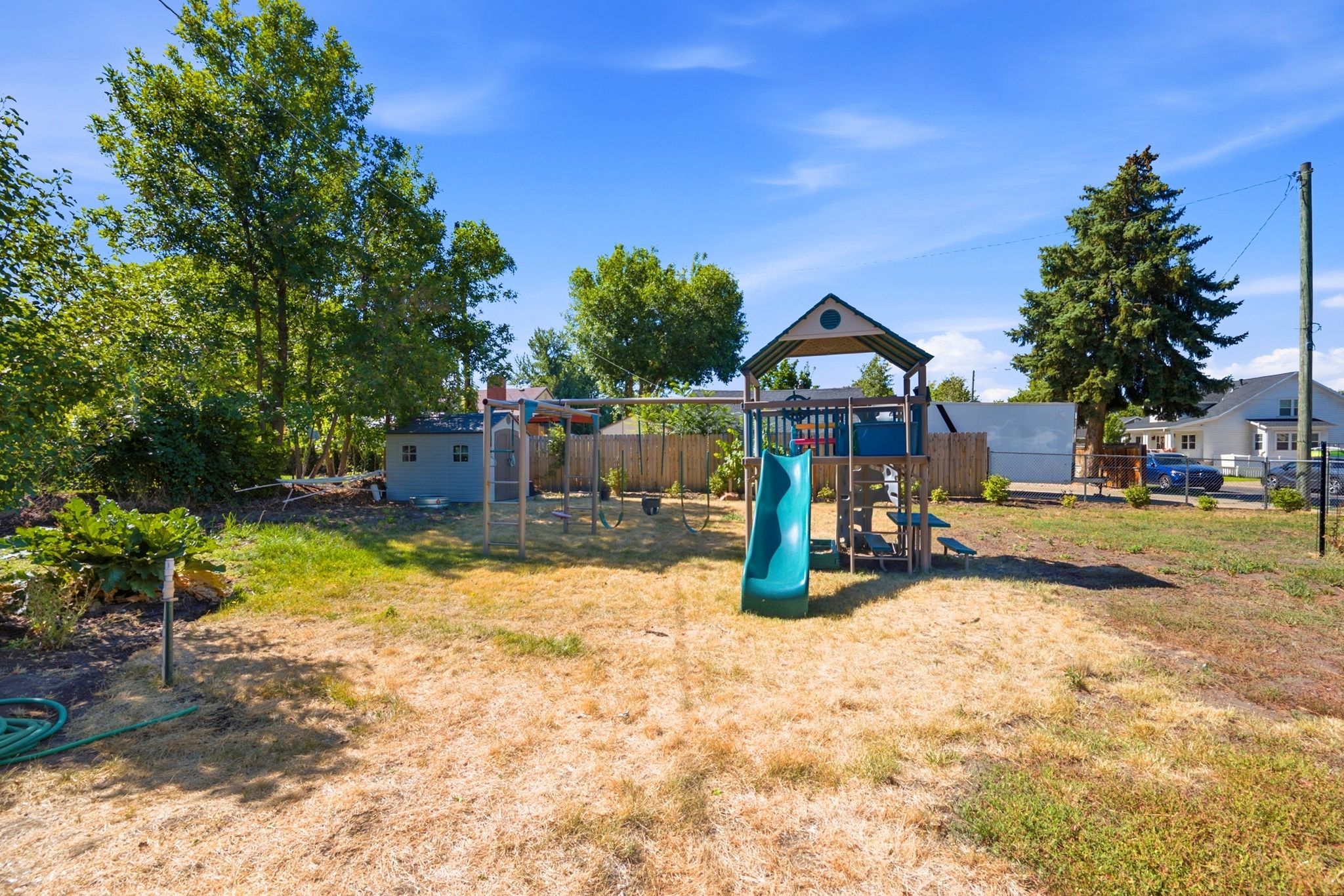 Spacious backyard featuring a wooden play structure with a slide, a swing set, mature trees, and a storage shed