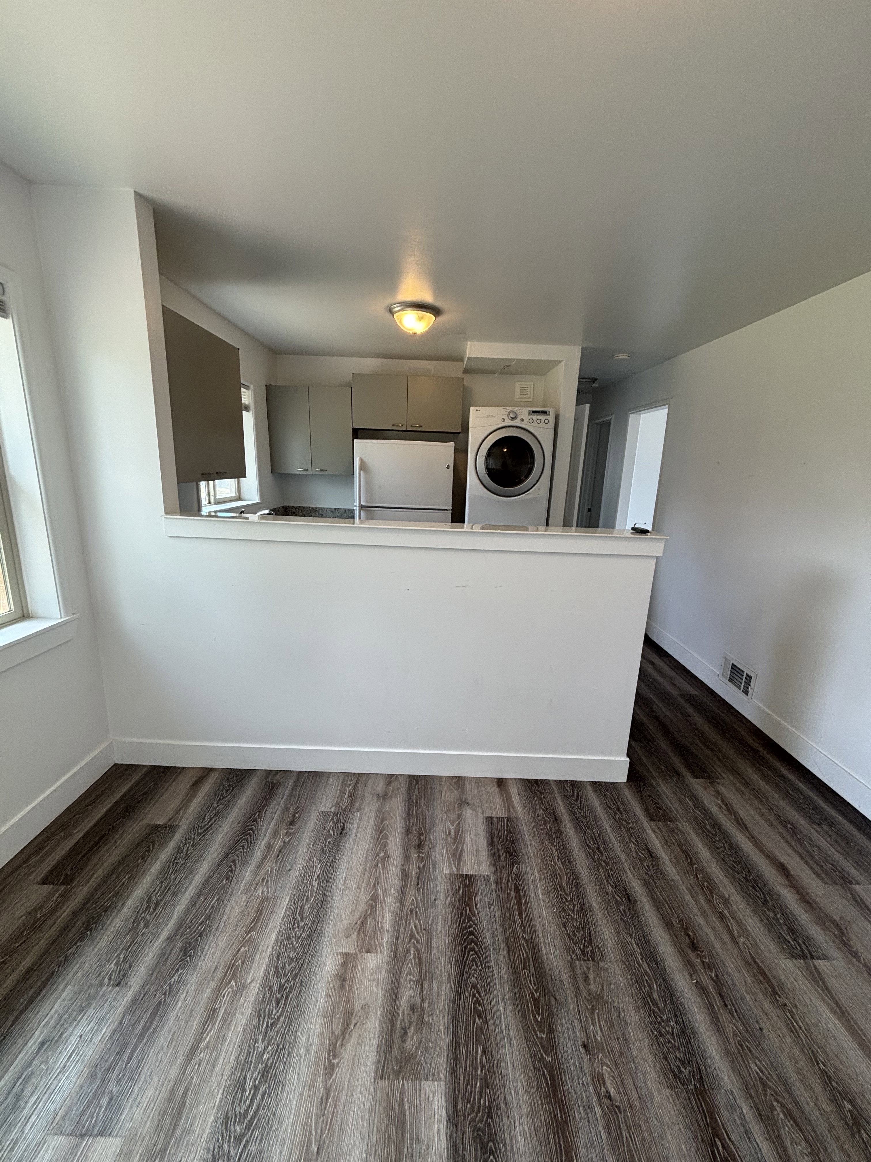 Open-concept living area featuring wood-finish flooring, a kitchen with light-colored cabinetry, a refrigerator, and a built-in washing machine/dryer.