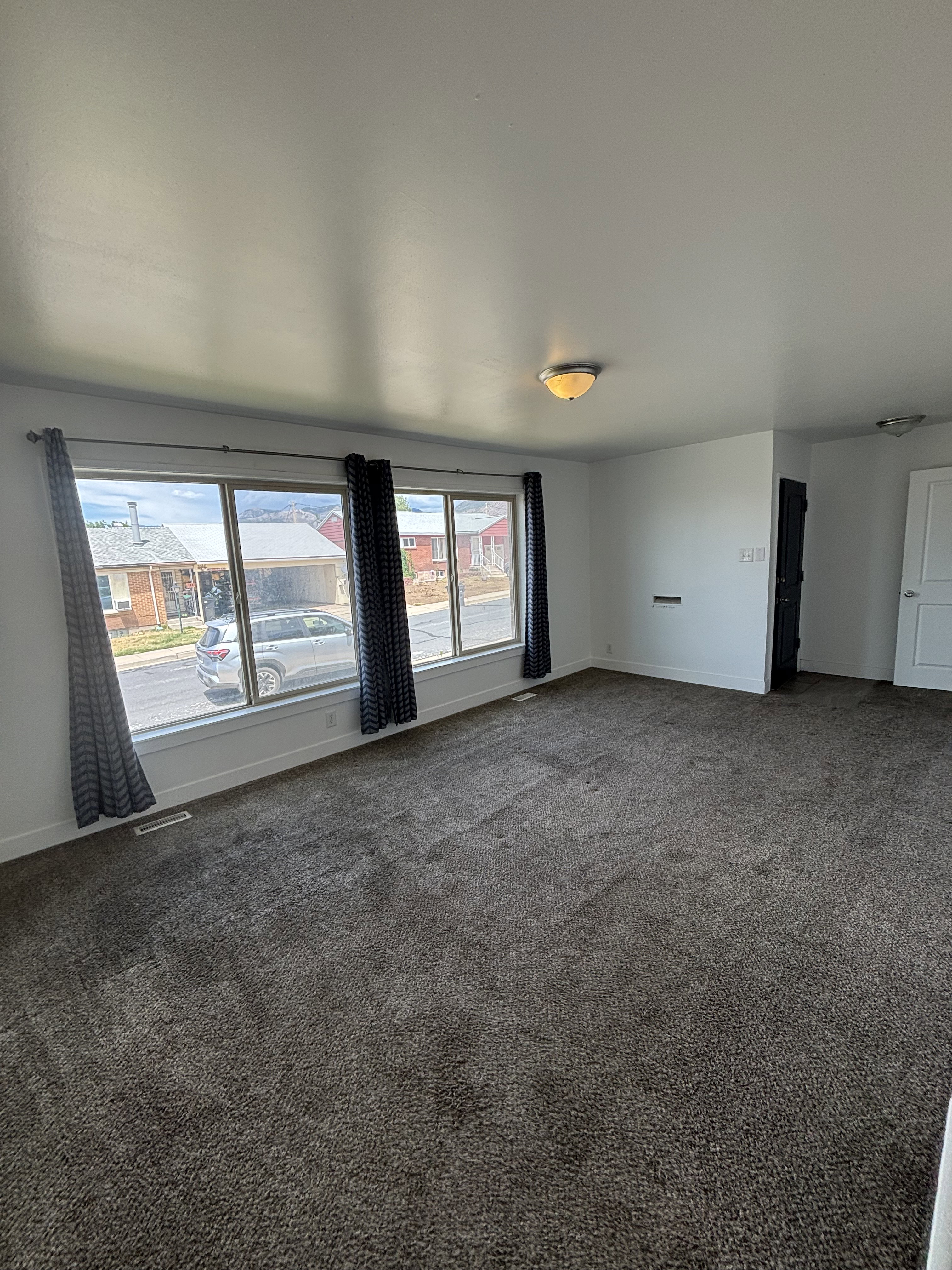 Spacious room featuring broad windows with white trim, dark patterned curtains, and plush dark gray carpeting.