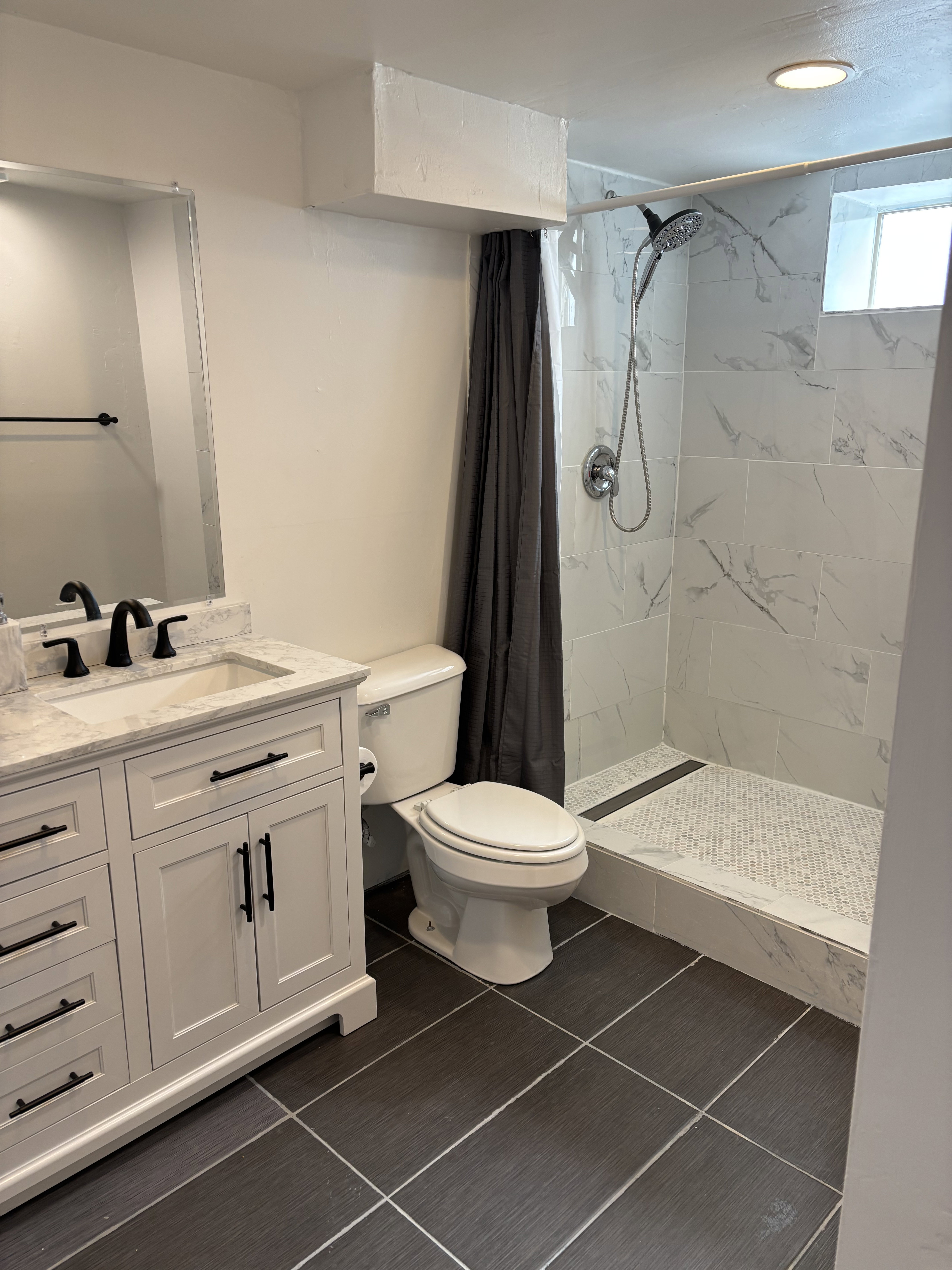 Modern bathroom featuring a white vanity with a marble-finish countertop, dark tile flooring, a walk-in shower with large format marble-finish wall tiles, a mosaic tile shower floor, and a contemporary shower head.