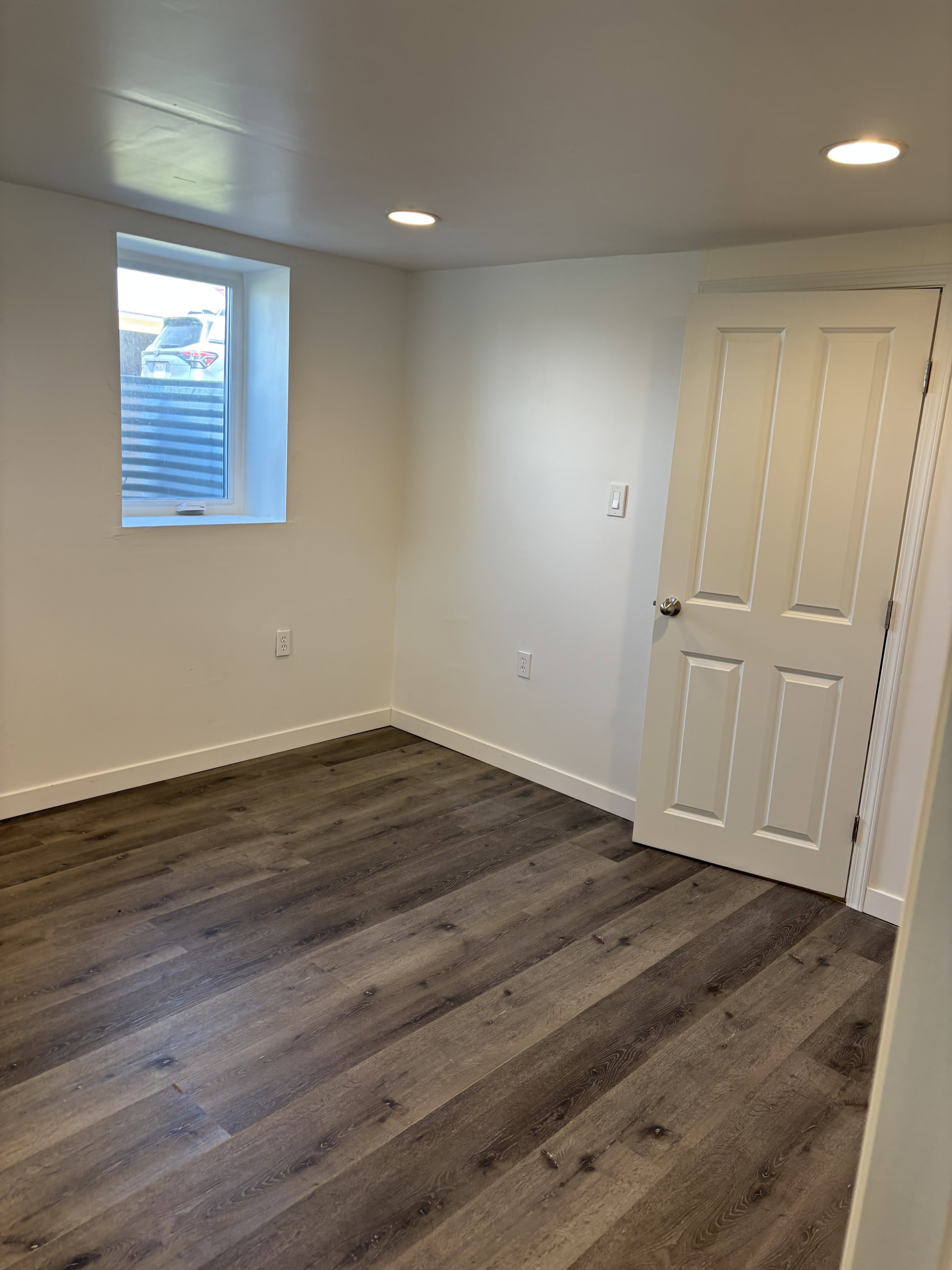 Interior room featuring dark wood-finish flooring, recessed ceiling lighting, a white painted door with a silver-tone handle, and a single window with horizontal blinds