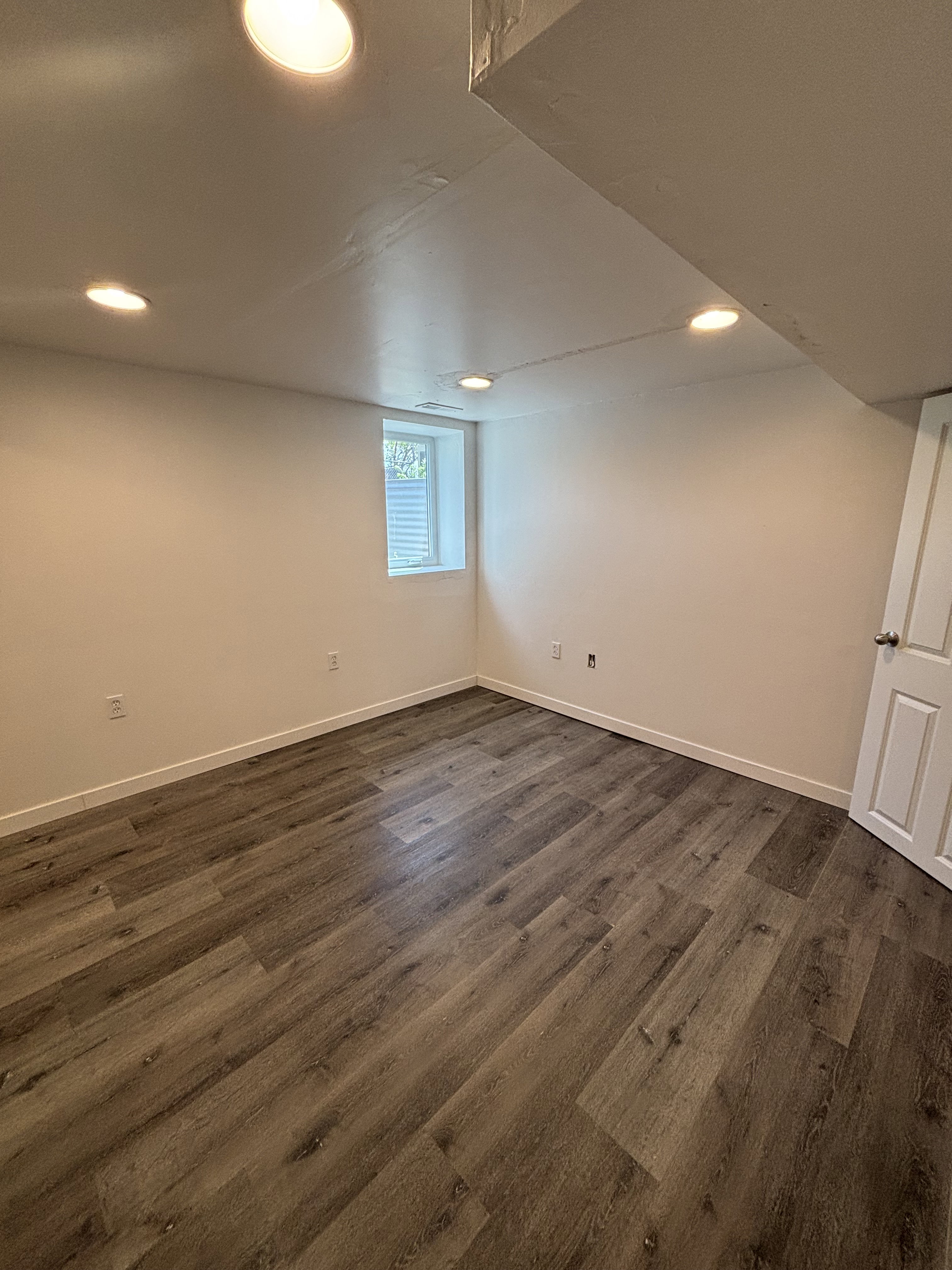 Spacious room featuring wood-finish flooring, white walls, recessed ceiling lighting, and a white interior door.