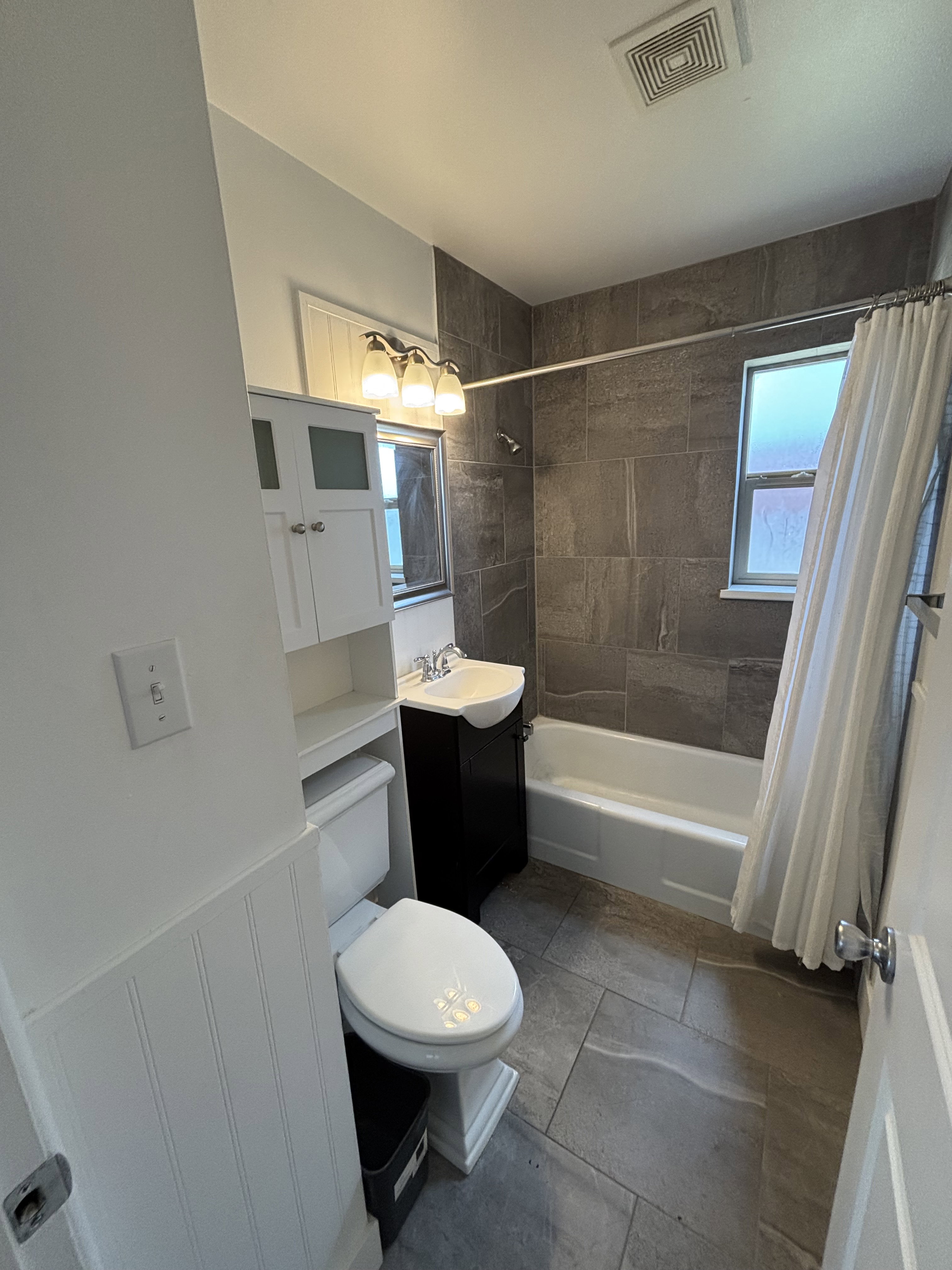 Bathroom featuring a shower-tub combination with dark tile surround, a dark wood-finish vanity with an integrated sink, and a white toilet.