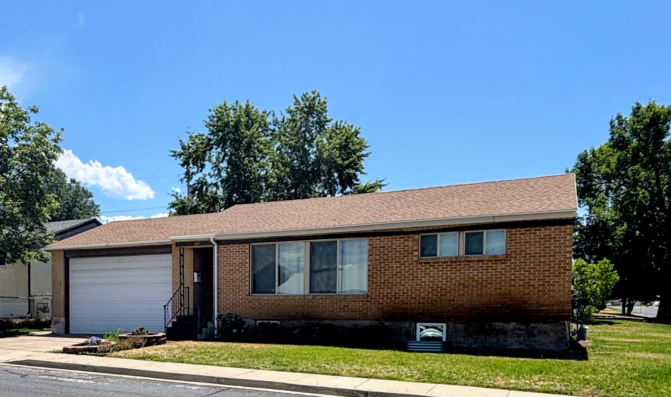Brick-front residence featuring an attached garage with a white door, a sloped shingle roof, and a mature tree-lined perimeter.