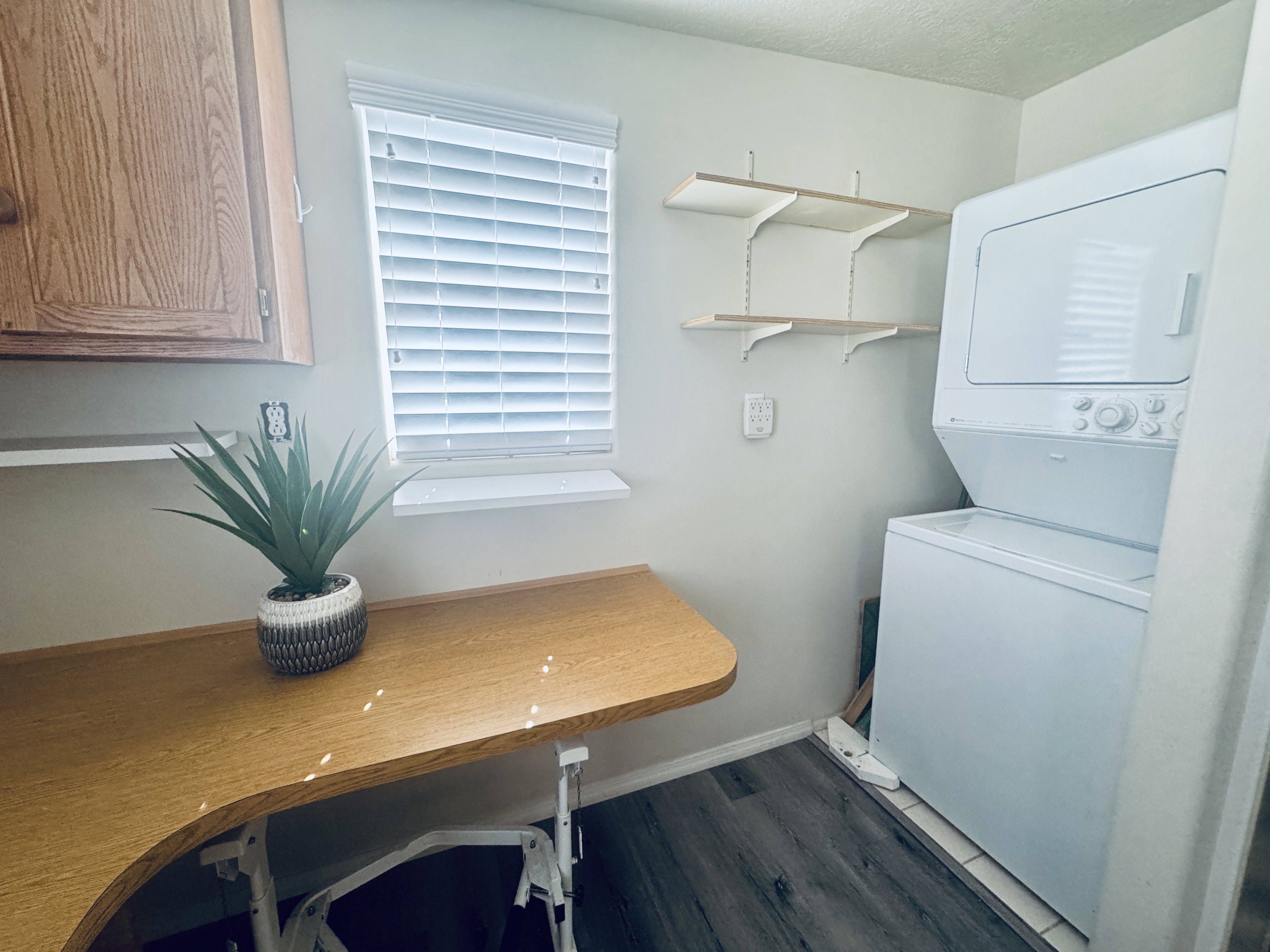 Dedicated laundry area featuring a stacked washer and dryer, wood-finish flooring, a window with horizontal blinds, built-in cabinetry, and wall-mounted shelving