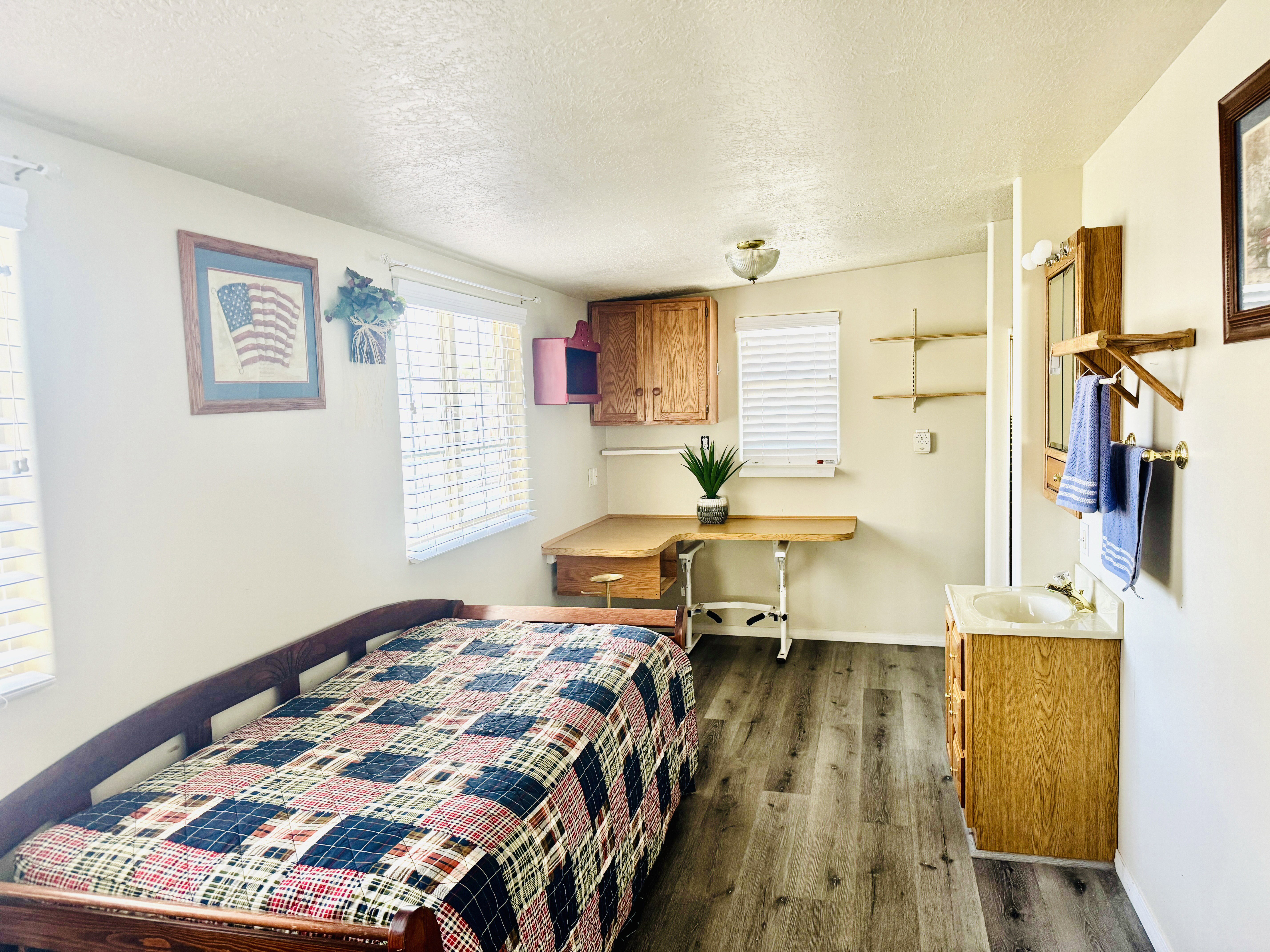 Spacious room featuring wood-finish flooring, two windows with blinds, a built-in desk with cabinet storage, a wall-mounted vanity with a sink, and open shelving