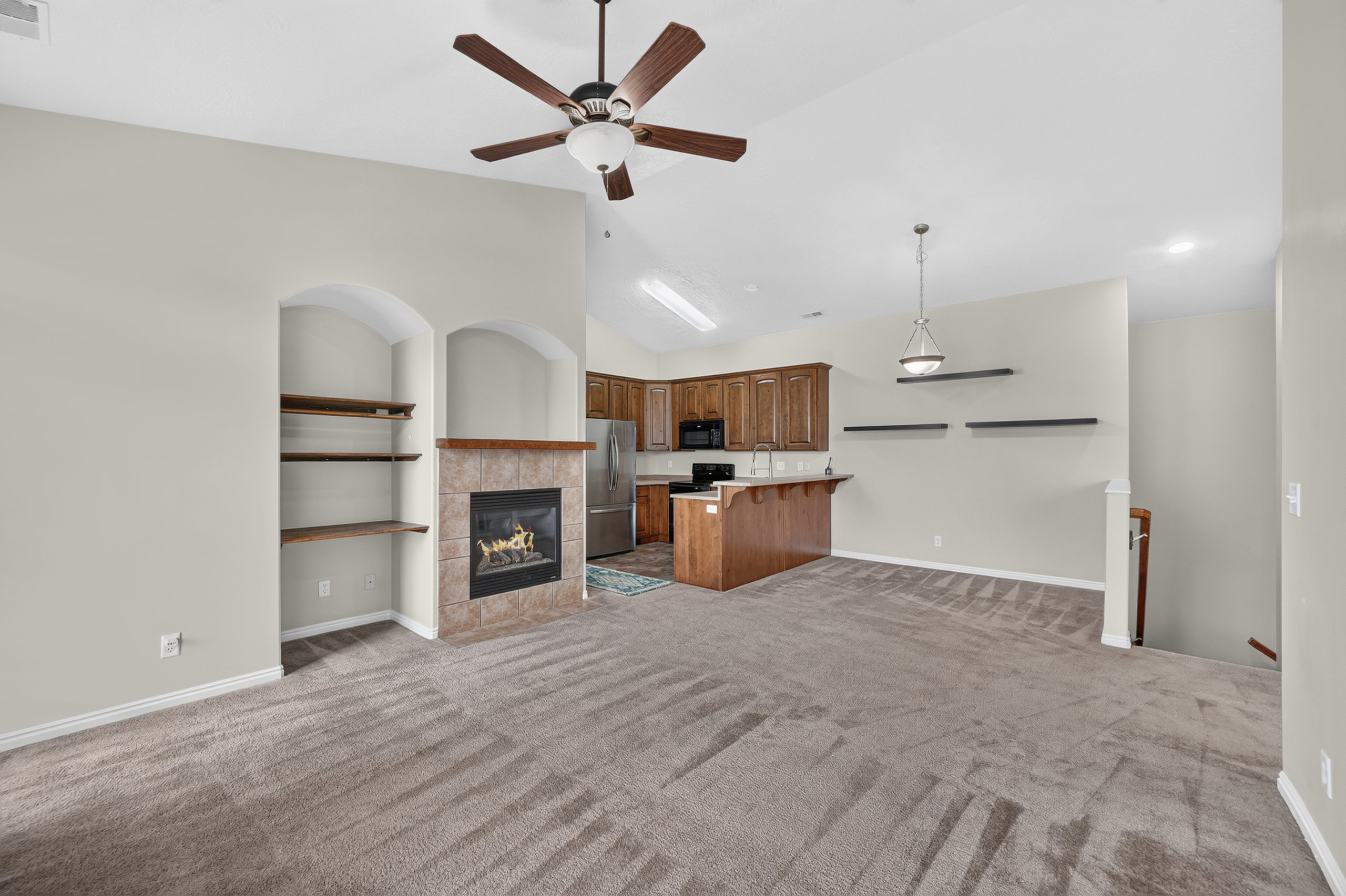 Living area featuring a vaulted ceiling with a ceiling fan, a tiled fireplace with a wood mantle, built-in arched shelving, and an open layout to the kitchen with wood cabinetry