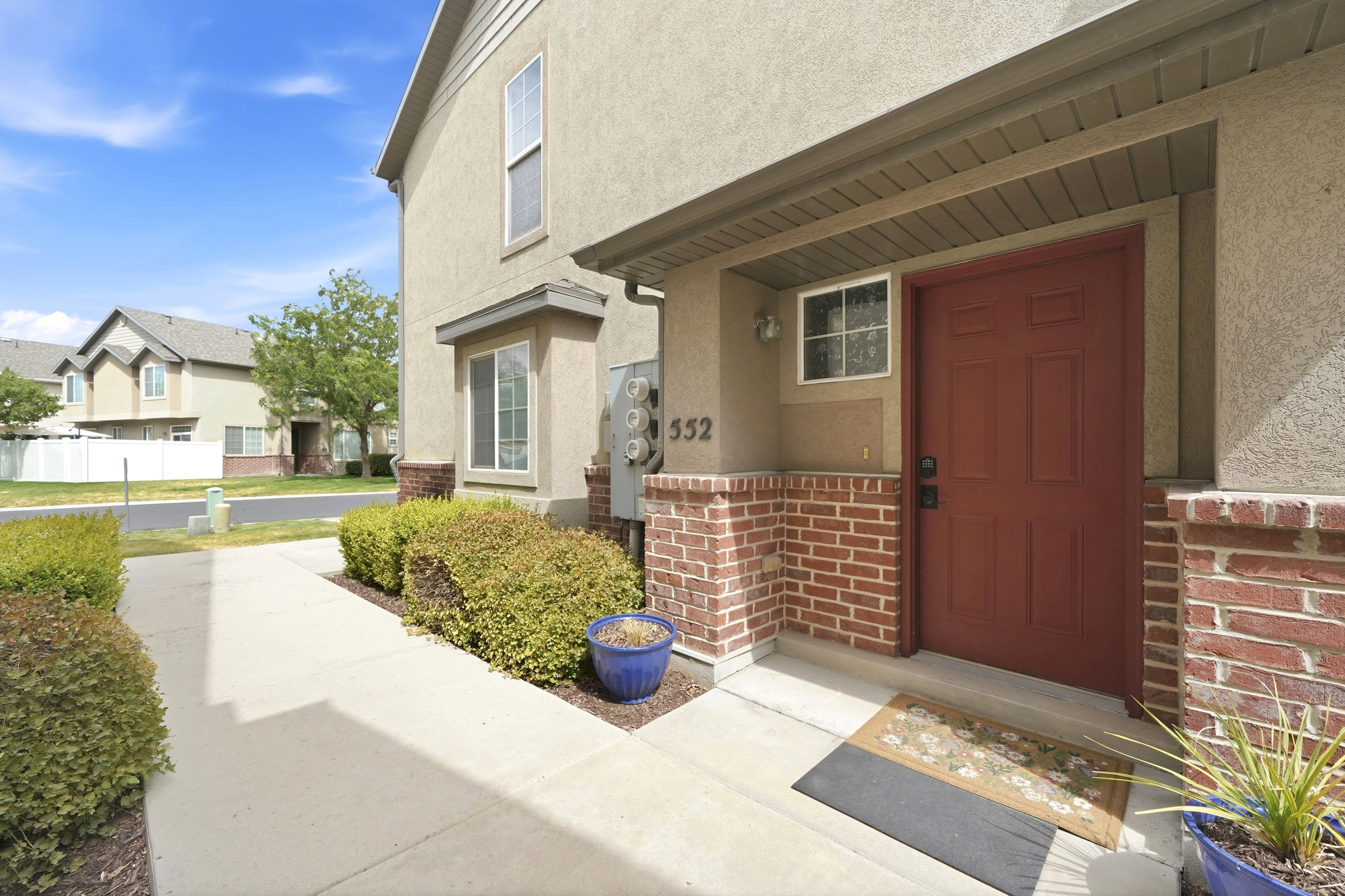 Entryway featuring a red paneled door, brick column accents, stucco exterior, covered porch, and concrete pathway