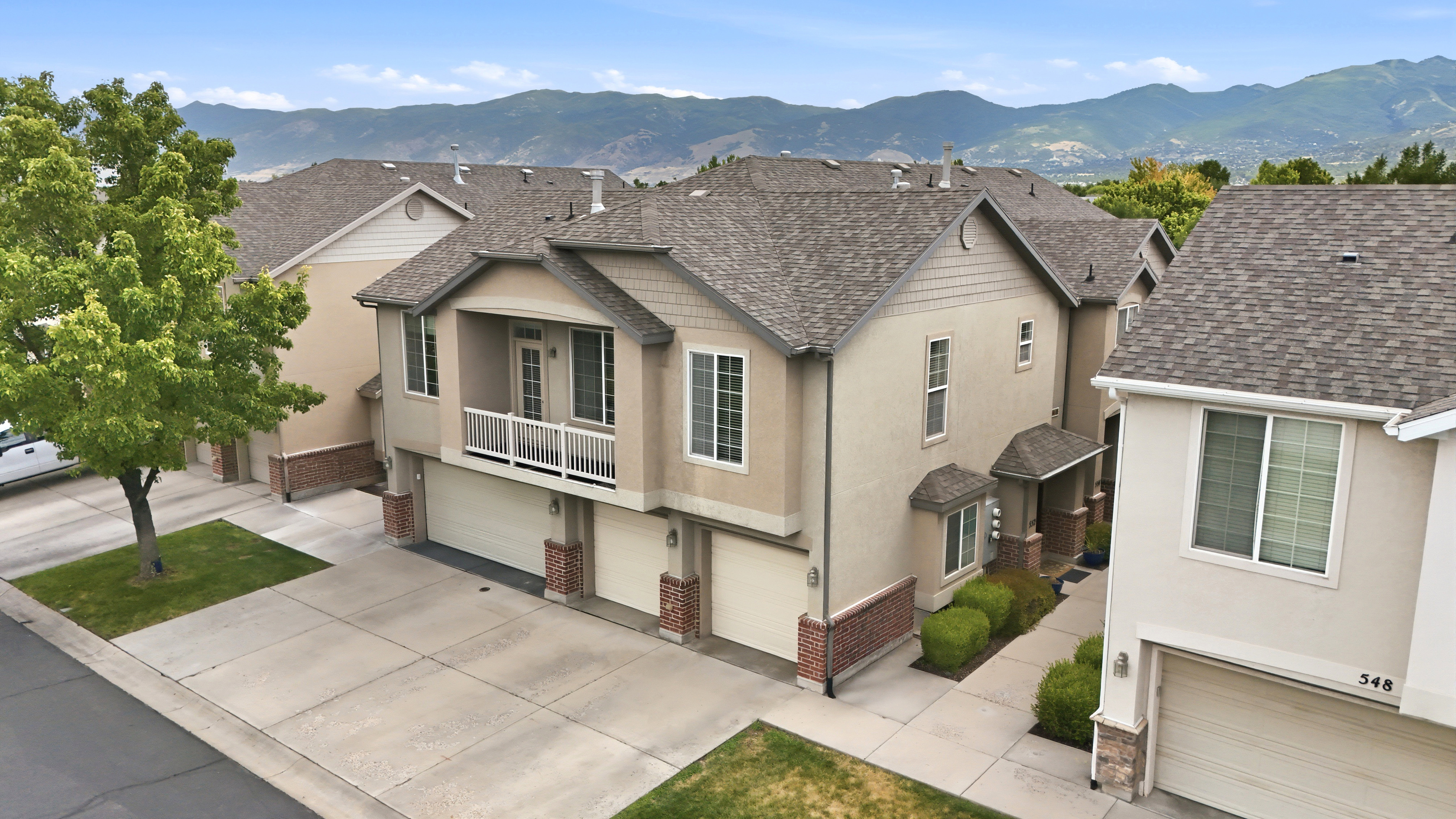 Multi-level townhome featuring a neutral stucco exterior with brick accents, attached two-car garage, and a second-story balcony with white railing