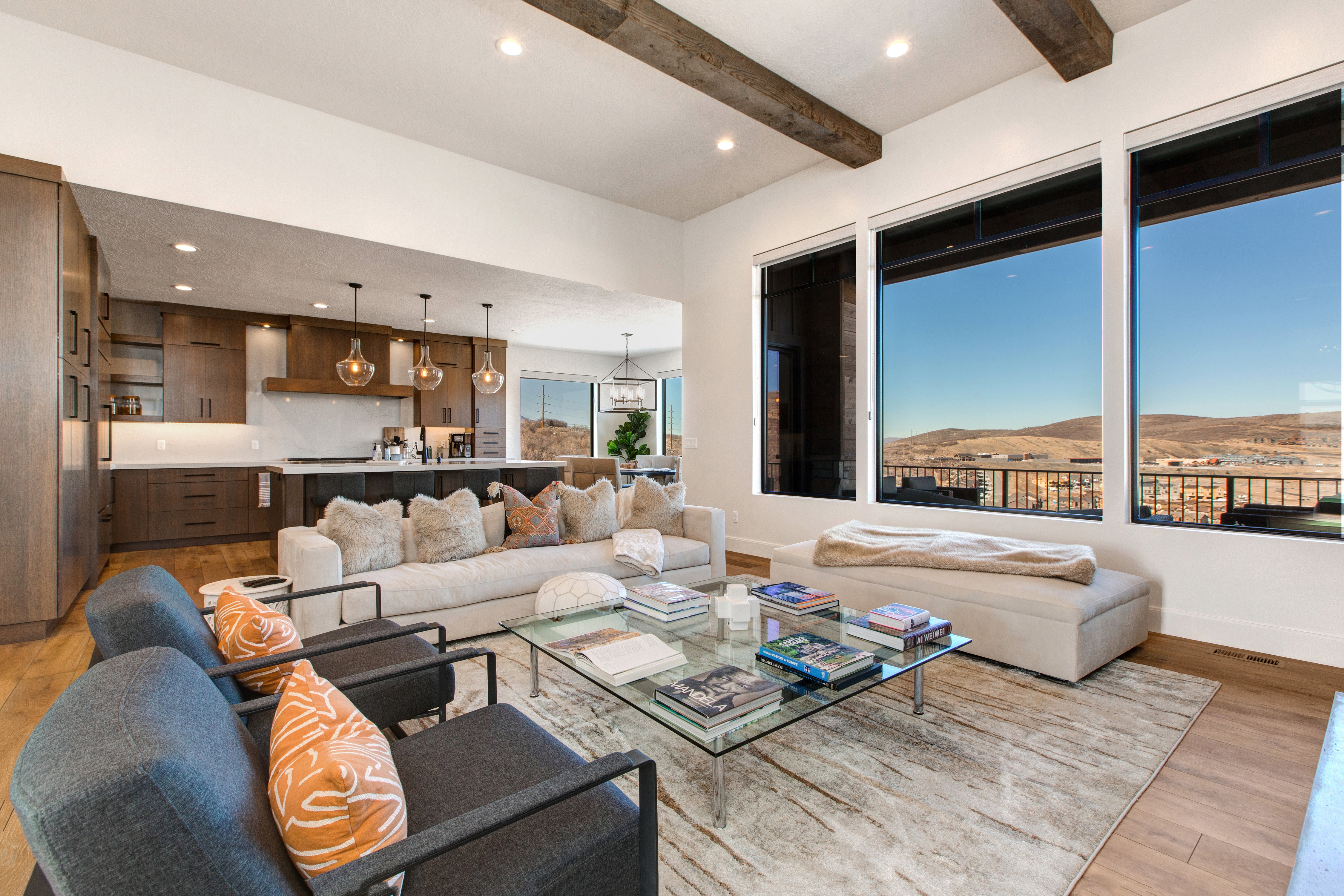 Open-concept living space featuring high ceilings with exposed wood beams, a modern kitchen with dark wood cabinetry and a large island, and expansive windows showcasing distant hills