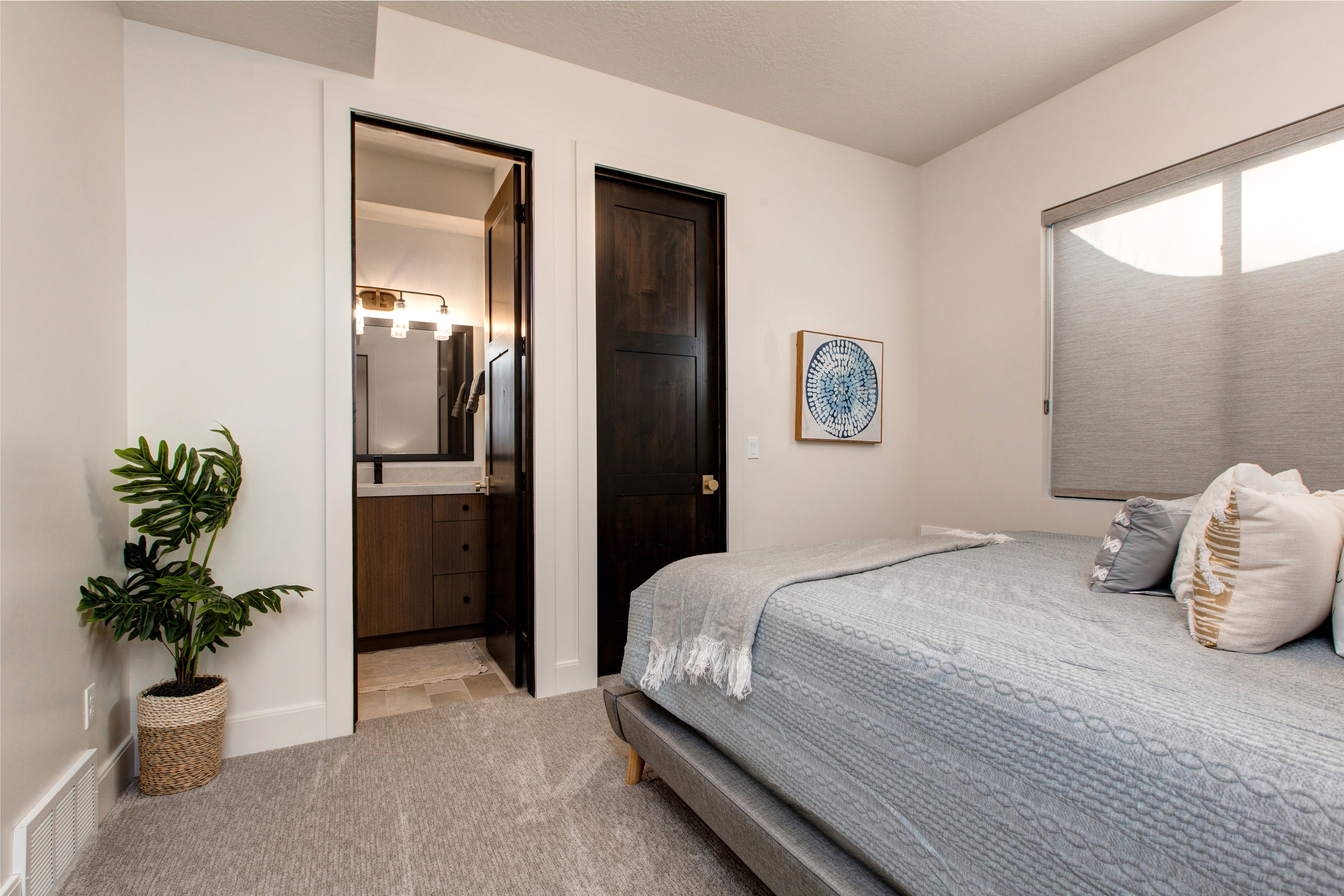 Carpeted bedroom featuring a dark wood-finish door, a window with a cellular shade, and a doorway opening to a bathroom with a wood-finish vanity and a wall-mounted light fixture