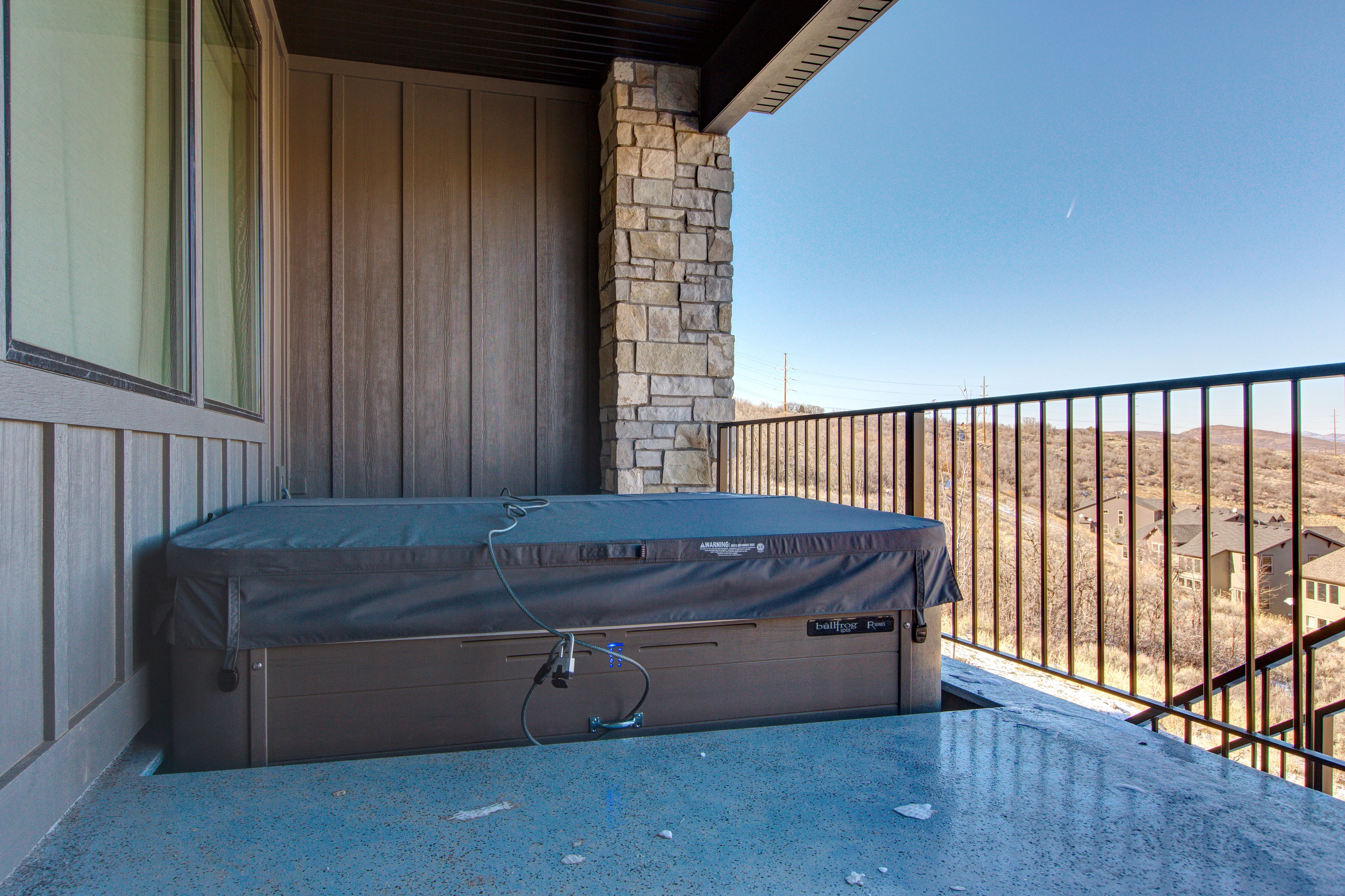 Outdoor hot tub on a concrete patio with a stone pillar, vertical panel siding, and black metal railing