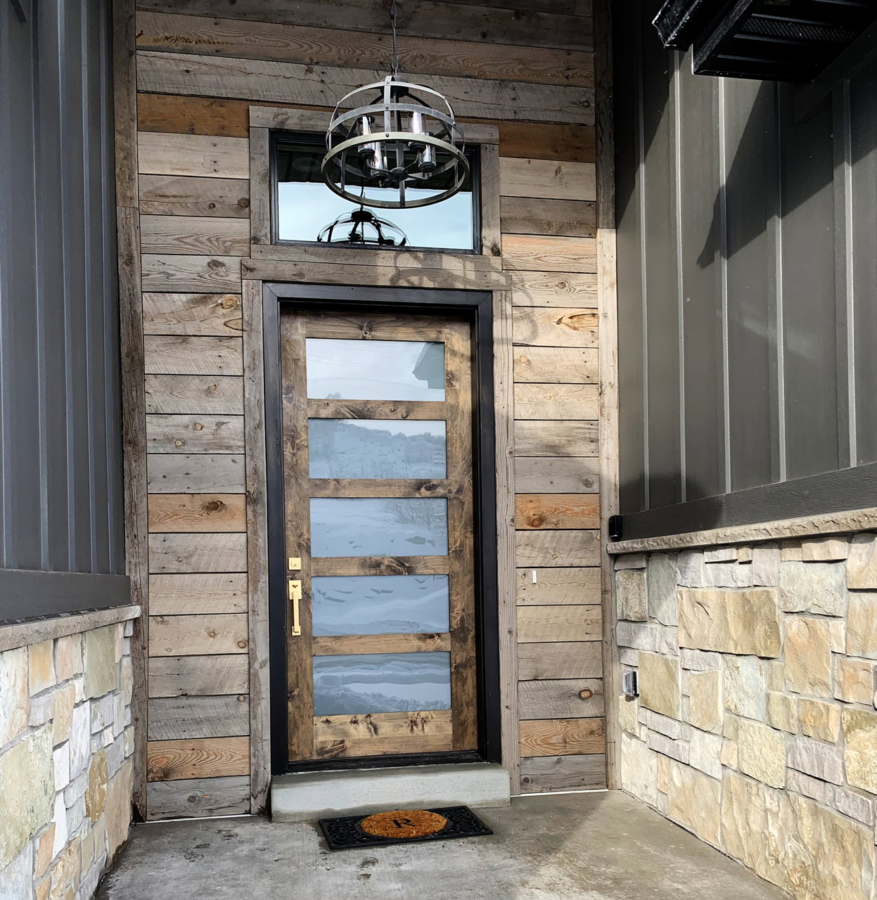 Modern entry featuring a five-panel glass door with a brass handle, rustic wood-finish siding, and a stone veneer foundation