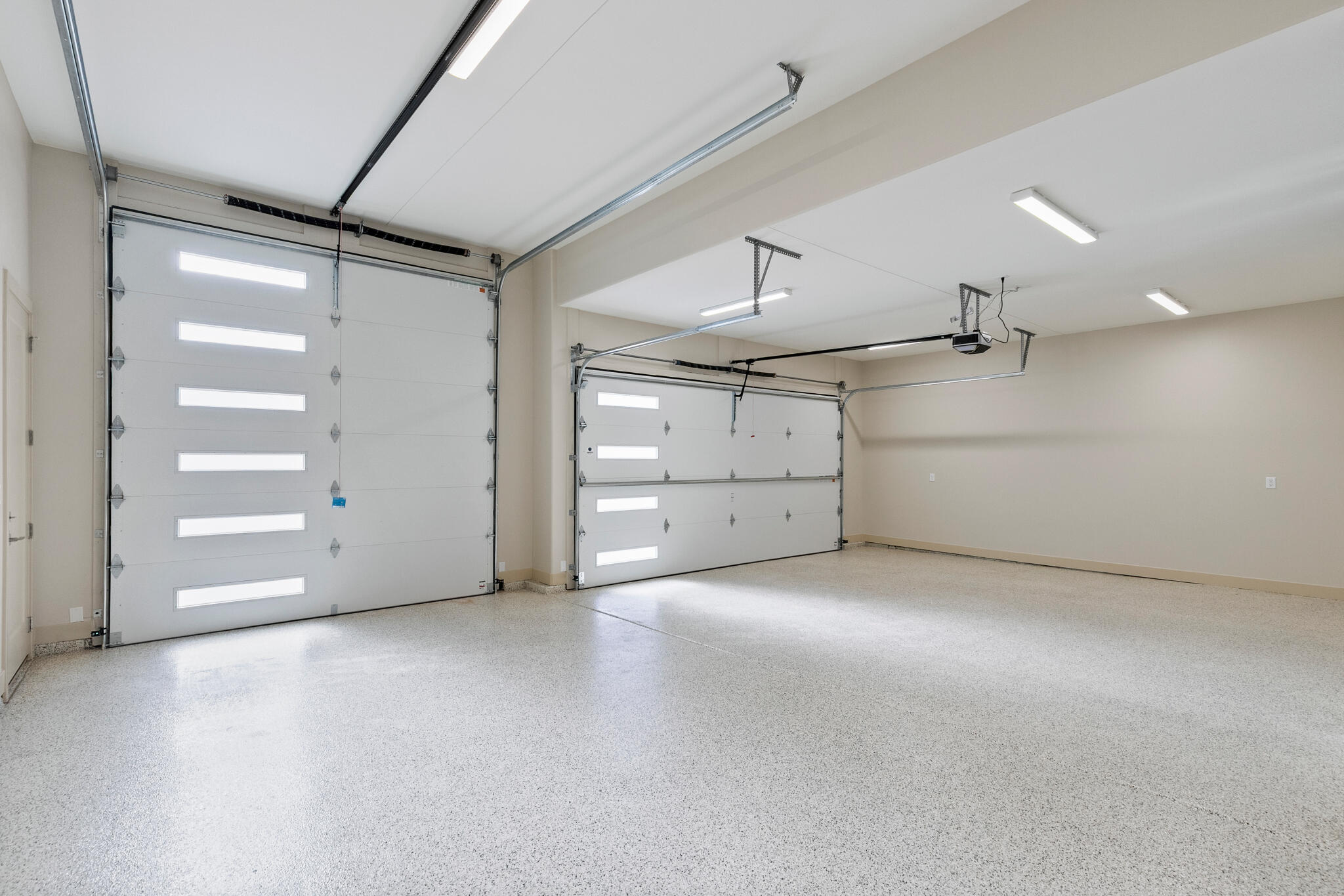Spacious garage featuring two overhead doors with integrated light panels, an epoxy-coated concrete floor, and multiple ceiling-mounted fluorescent light fixtures