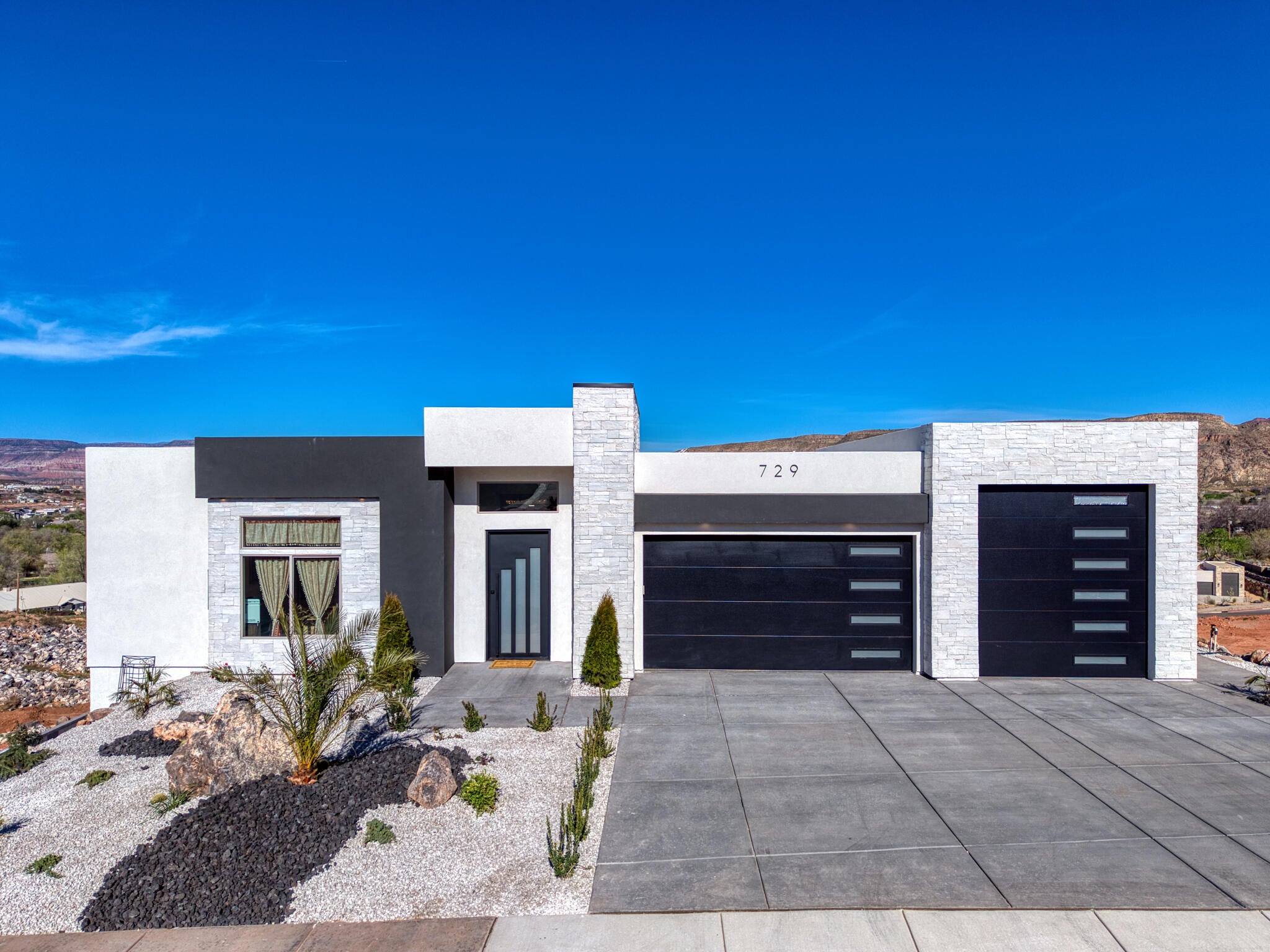 Modern exterior featuring a prominent black and white facade, stacked stone accents, a dark entry door with vertical glass inserts, and two black garage doors with horizontal window panels