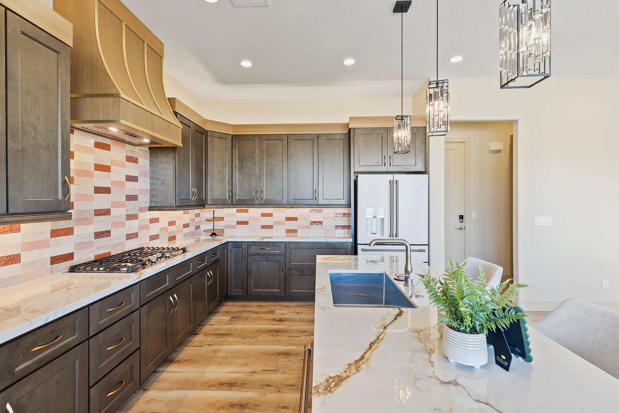 Spacious kitchen featuring an extended island with a built-in sink, stone countertops, wood-finish flooring, a decorative tile backsplash, and dark cabinetry with brass-finish hardware