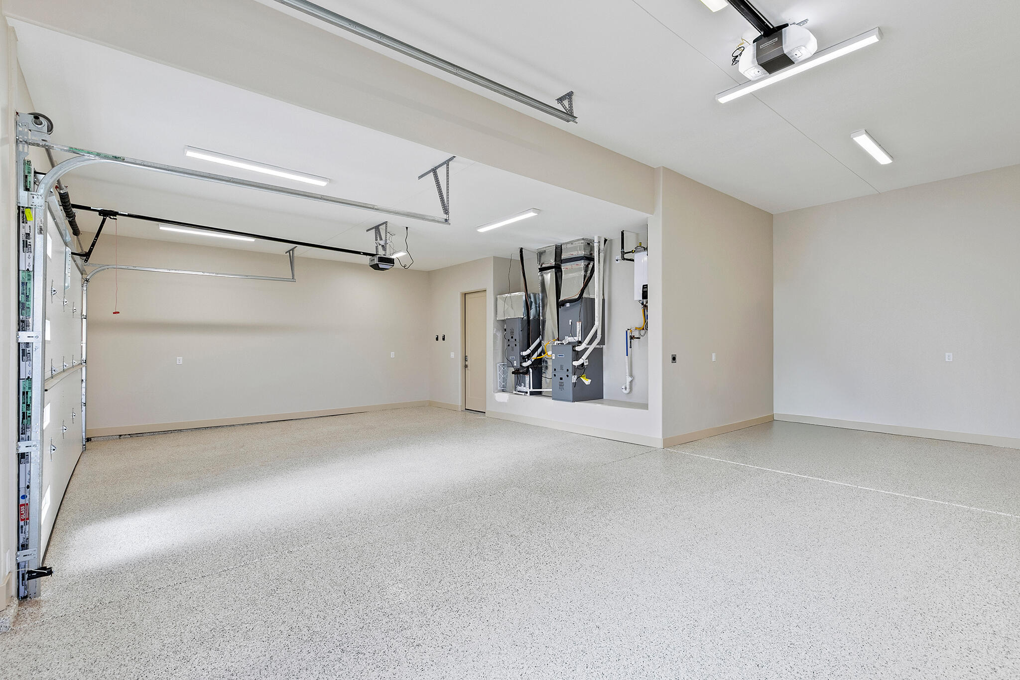 Spacious garage featuring a durable epoxy floor coating, multiple overhead lighting fixtures, exposed garage door mechanisms, and a utility area with visible mechanical systems