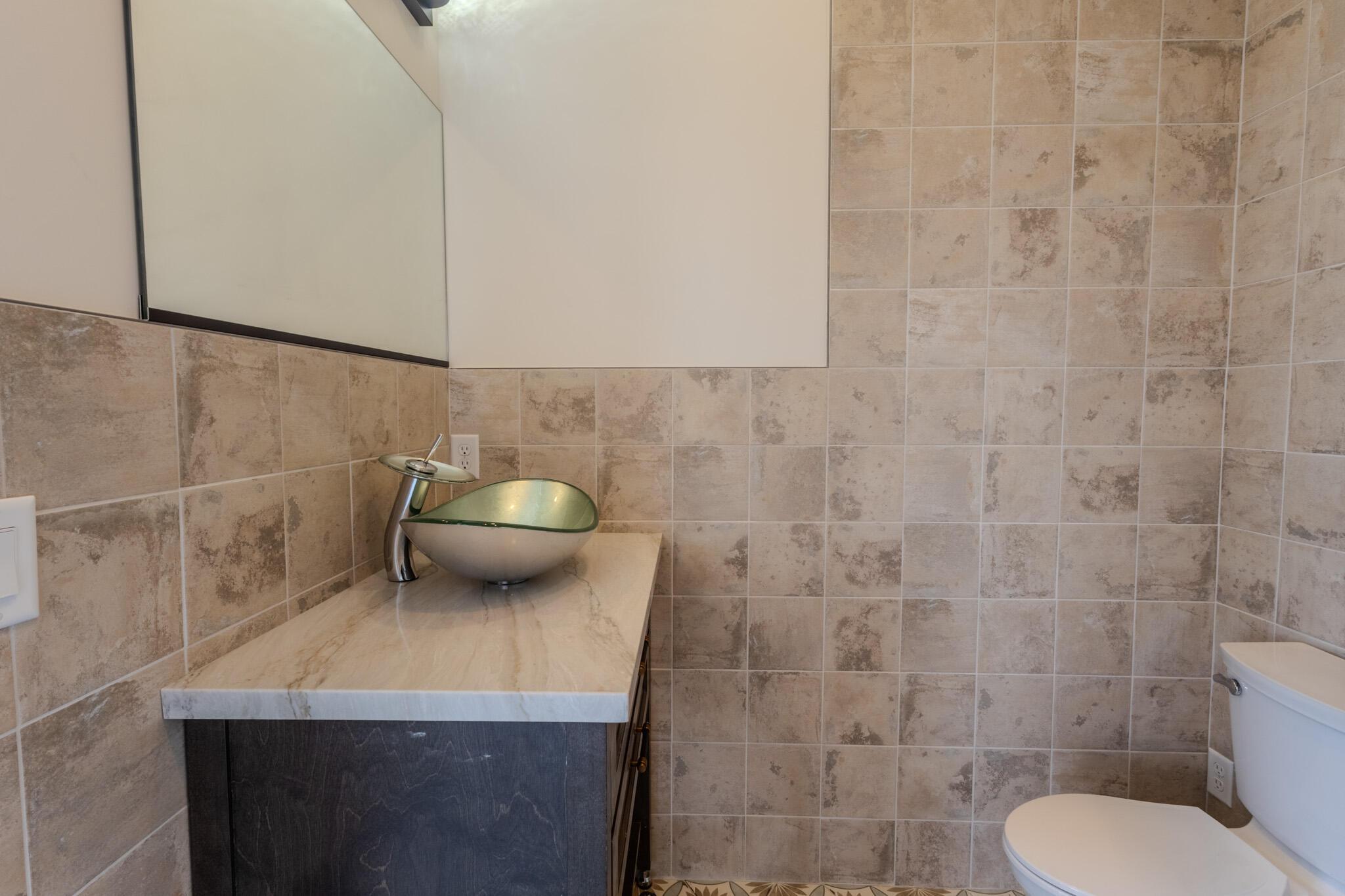 Bathroom featuring a vessel sink with a contemporary faucet, a dark wood-finish vanity, a light-toned countertop, and tiled walls extending to the floor