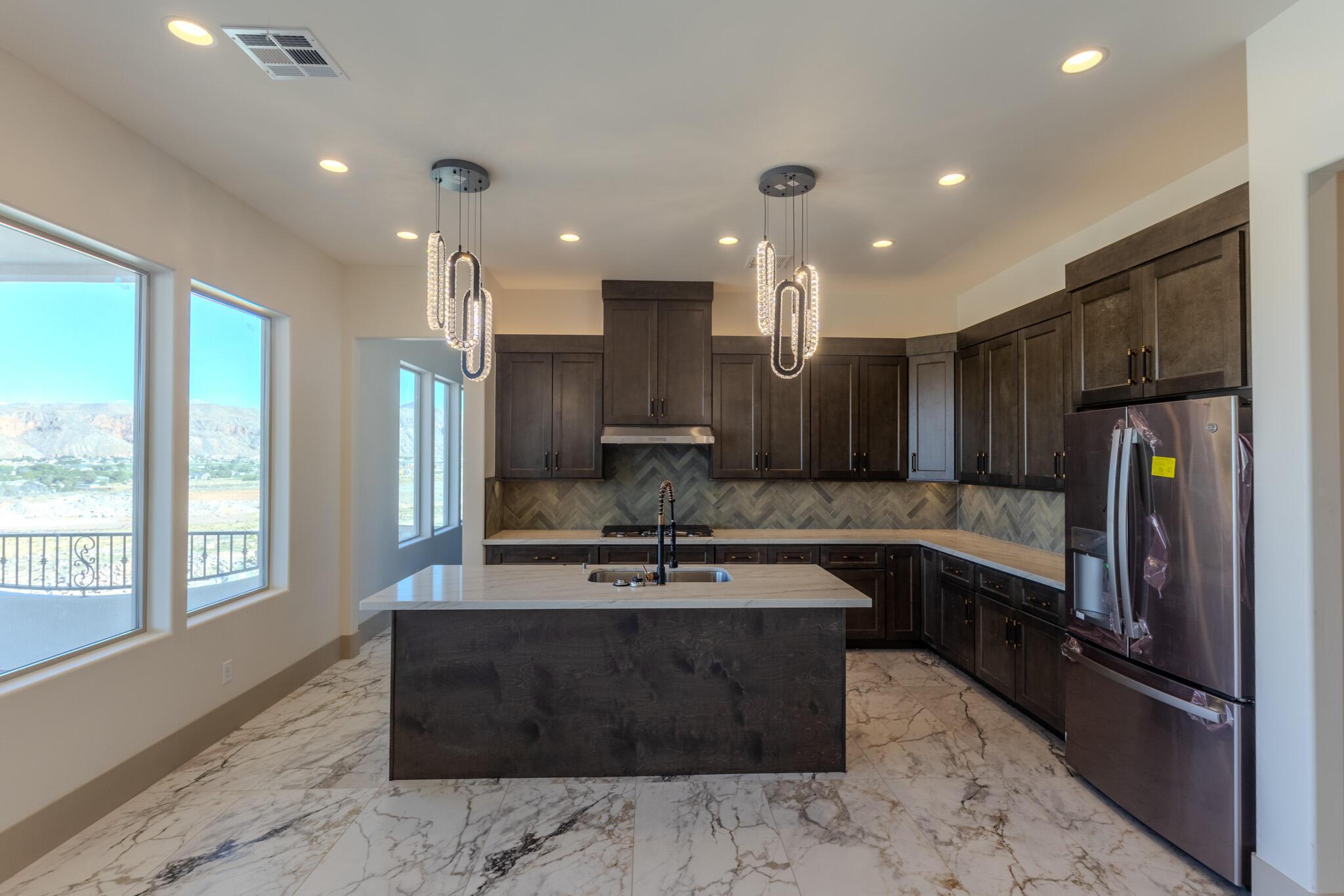 Contemporary kitchen featuring a central island with a sink, pendant lighting, dark wood cabinetry, stainless steel appliances, and large windows