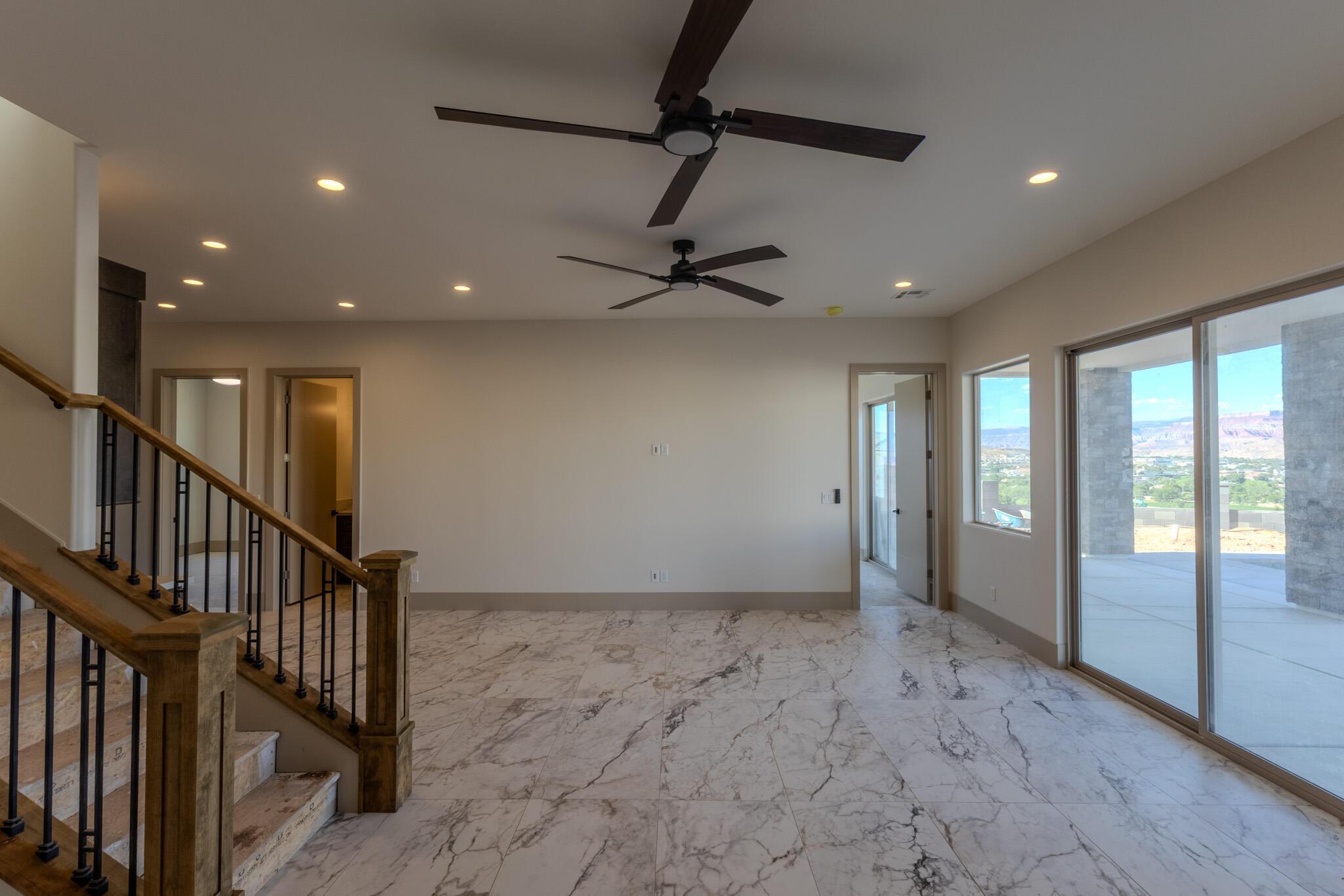Spacious room featuring elegant marble-finish flooring, two ceiling fans, recessed lighting, and a prominent staircase with wood handrails and black metal balusters