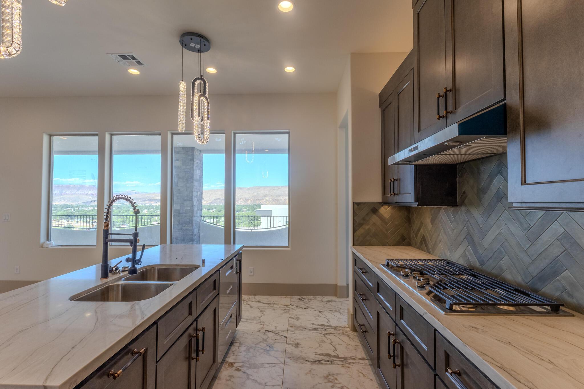 Modern kitchen featuring a large island with dual sinks, a gas cooktop with a stainless steel hood, dark wood-finish cabinetry, light-toned countertops, and a herringbone pattern tile backsplash