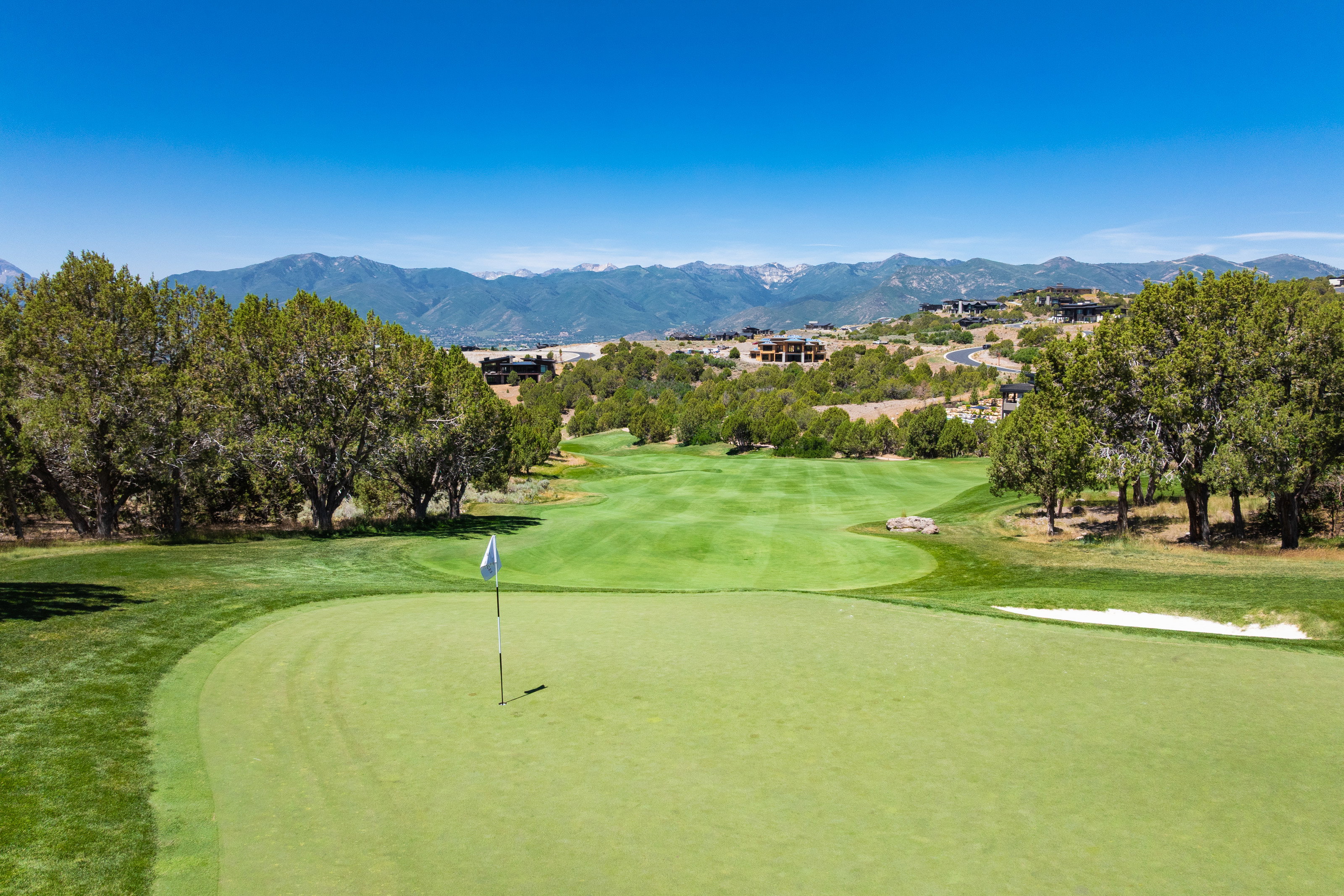 Expansive golf course setting featuring a putting green with flagstick, surrounded by manicured fairways, mature trees, and distant mountain ranges