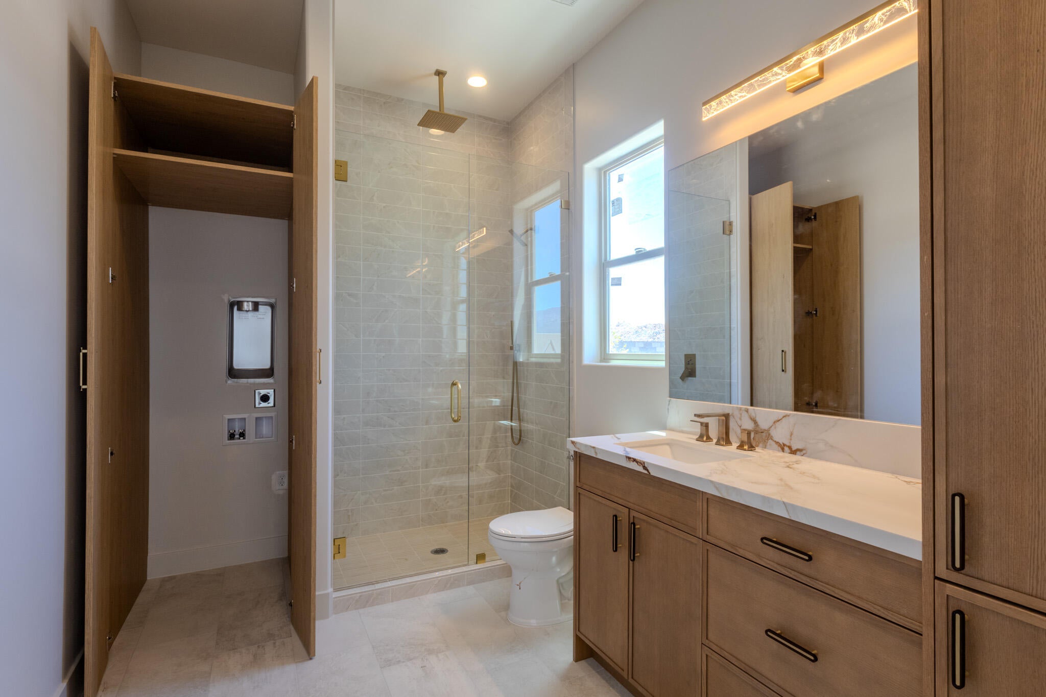 Contemporary bathroom featuring a spacious glass-enclosed shower with a rainfall showerhead, a single vanity with a stone-finish countertop and brass fixtures, and a concealed laundry area with hookups
