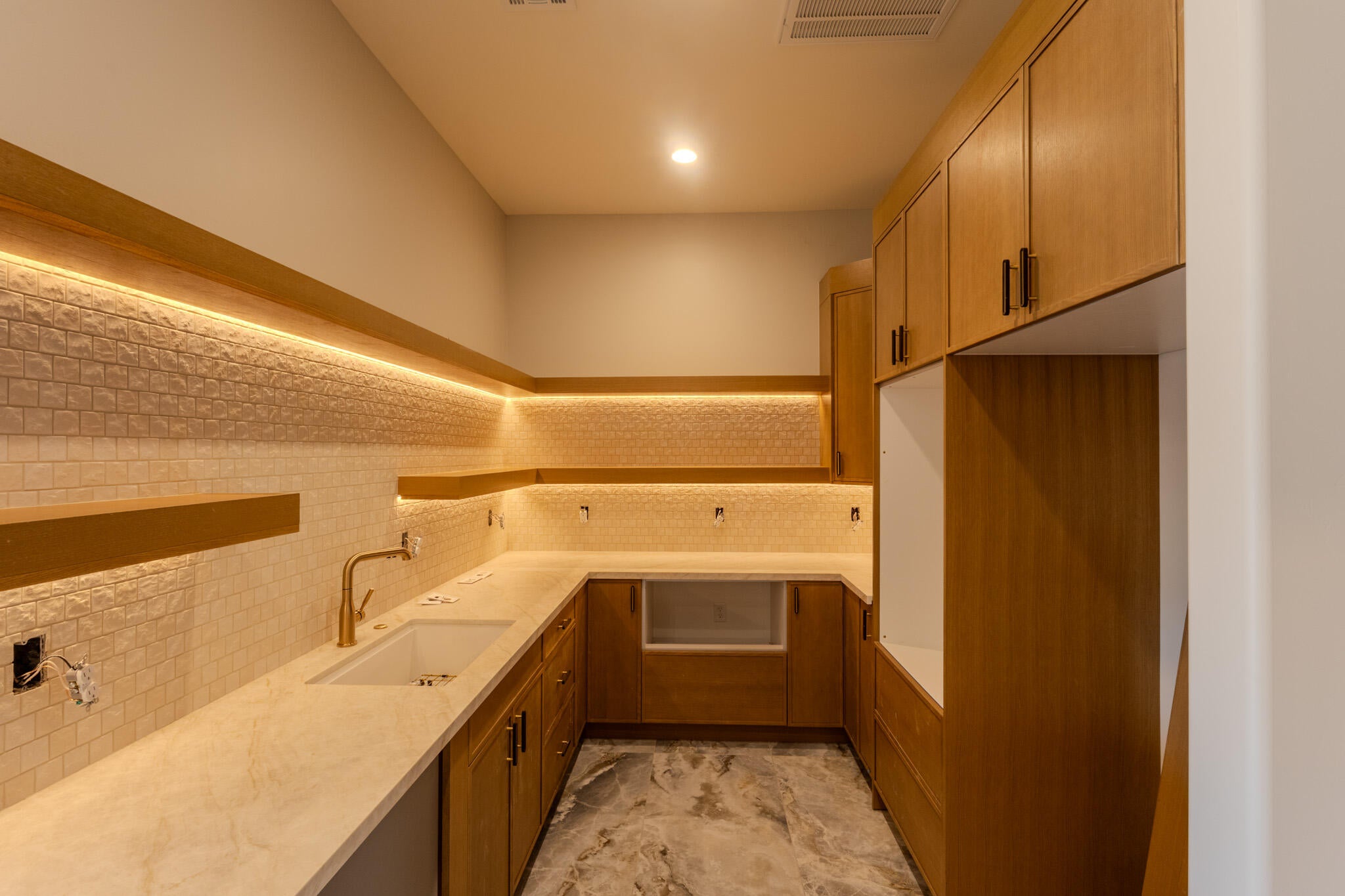 Modern kitchen space featuring extensive wood cabinetry with dark hardware, under-cabinet lighting, a textured tile backsplash, and light-toned countertops