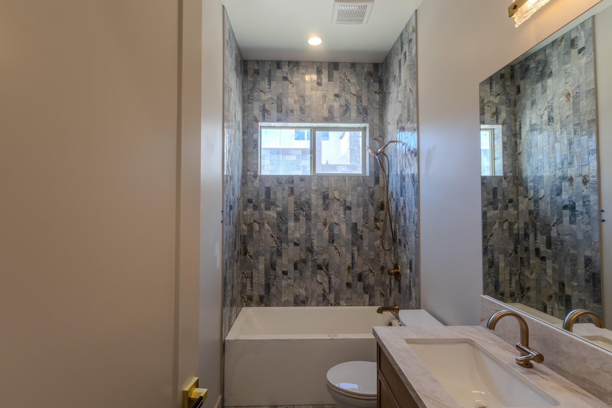 Contemporary bathroom featuring a tub-shower combination with floor-to-ceiling multi-tone tile surround, a built-in window, and a vanity with an integrated sink and bronze-finish hardware