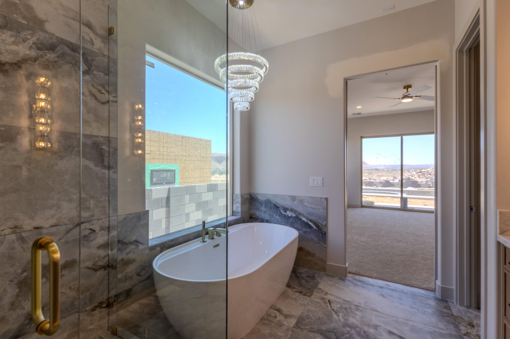 Freestanding soaking tub with floor-to-ceiling window, accompanied by a glass-enclosed shower with a gold-tone handle and integrated wall sconce lighting