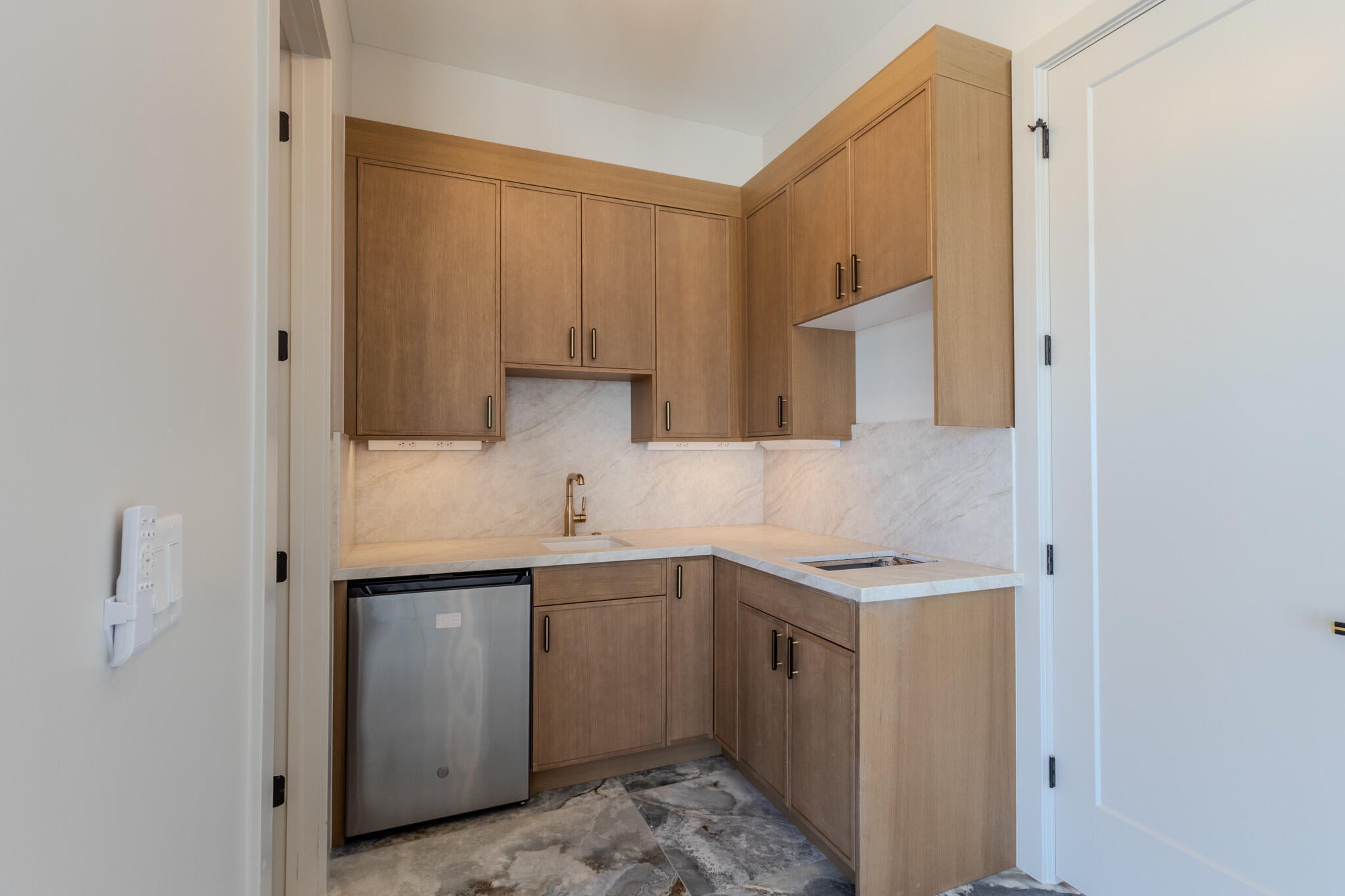Wet bar featuring light wood-finish cabinetry with modern hardware, under-cabinet lighting, a built-in stainless steel beverage refrigerator, and stone countertops with a matching backsplash