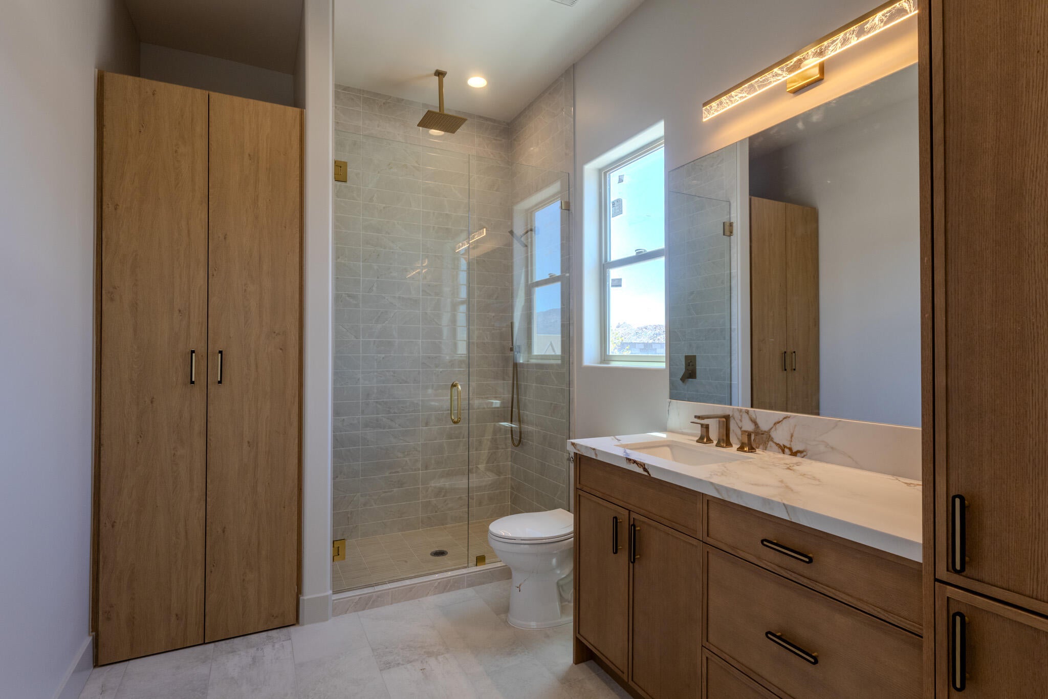 Bathroom featuring a spacious glass-enclosed shower with subway tile surround and rain shower head, a single vanity with stone countertop and integrated sink, coordinating wood-finish cabinetry, and a large mirror with integrated lighting
