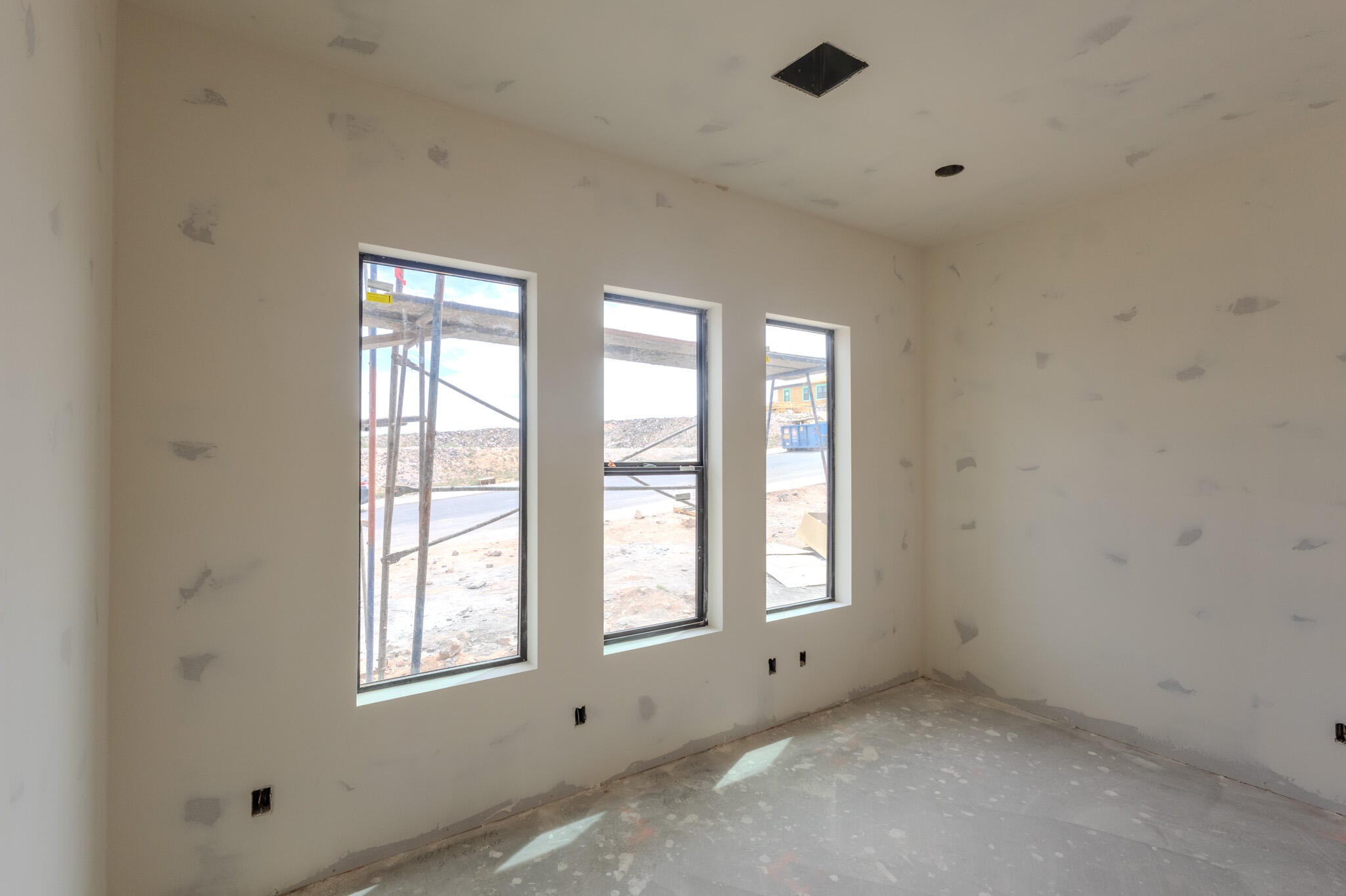 Room under construction featuring three vertical windows with black frames, white walls, exposed concrete flooring, a recessed ceiling light, and a square ceiling vent