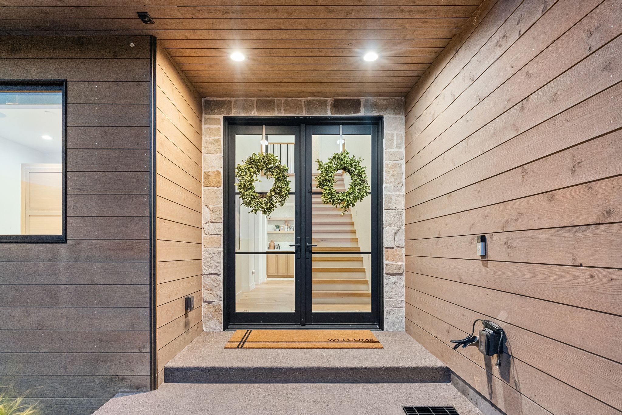 Modern architectural entrance featuring dark-framed double glass doors, stone facade accents, horizontal wood-finish siding, and a wood-panelled ceiling with recessed lighting