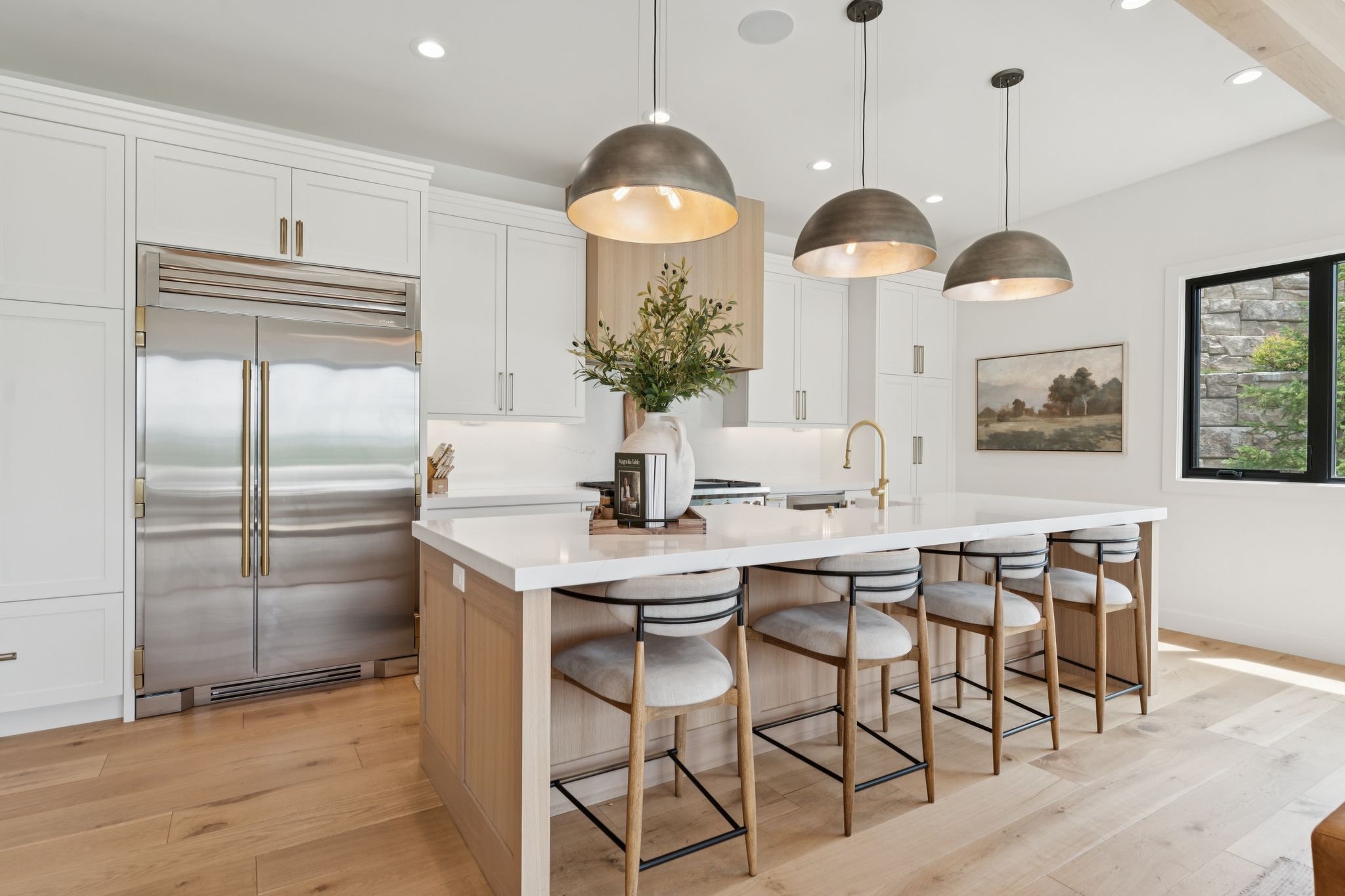 Gourmet kitchen featuring a large central island with seating, a stainless steel built-in refrigerator, light wood-finish flooring, white cabinetry with gold-tone hardware, and three dome pendant lights